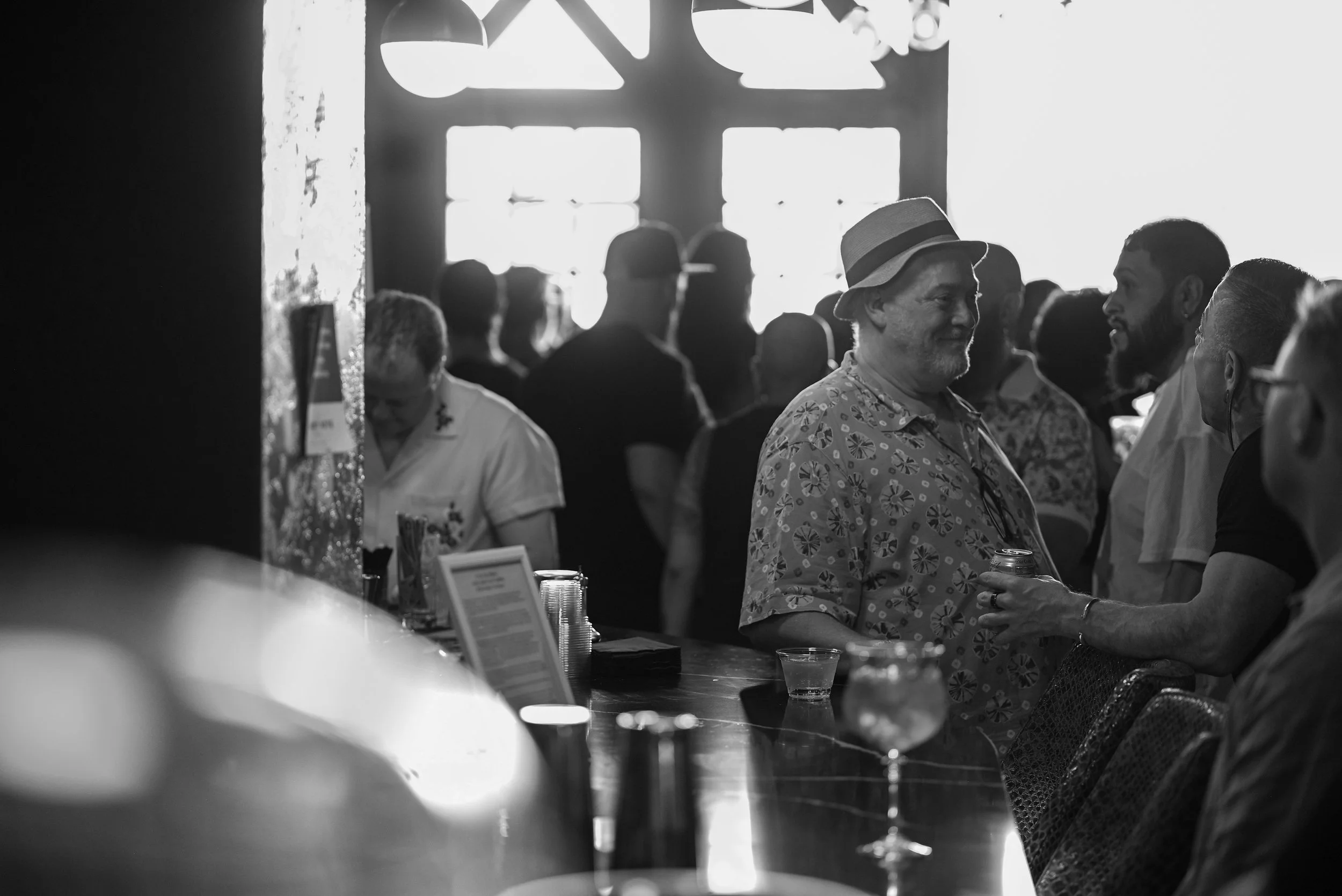 People socializing at a bar or event, with some engaged in conversation, in a room with large windows and hanging light fixtures, in black and white.