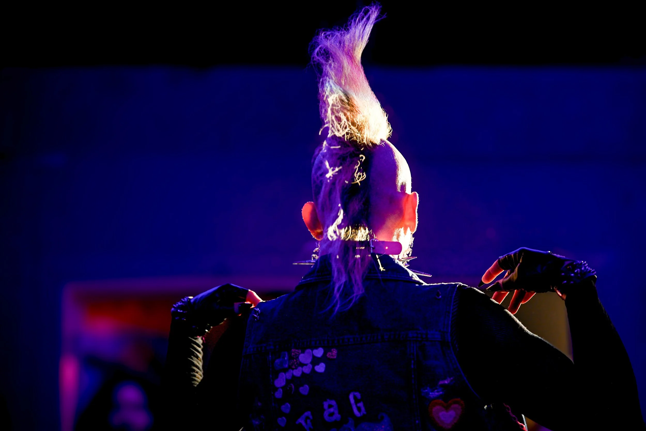 Person with long hair and dark outfit standing with arms outstretched in front of a dark background, illuminated by purple and blue lights.