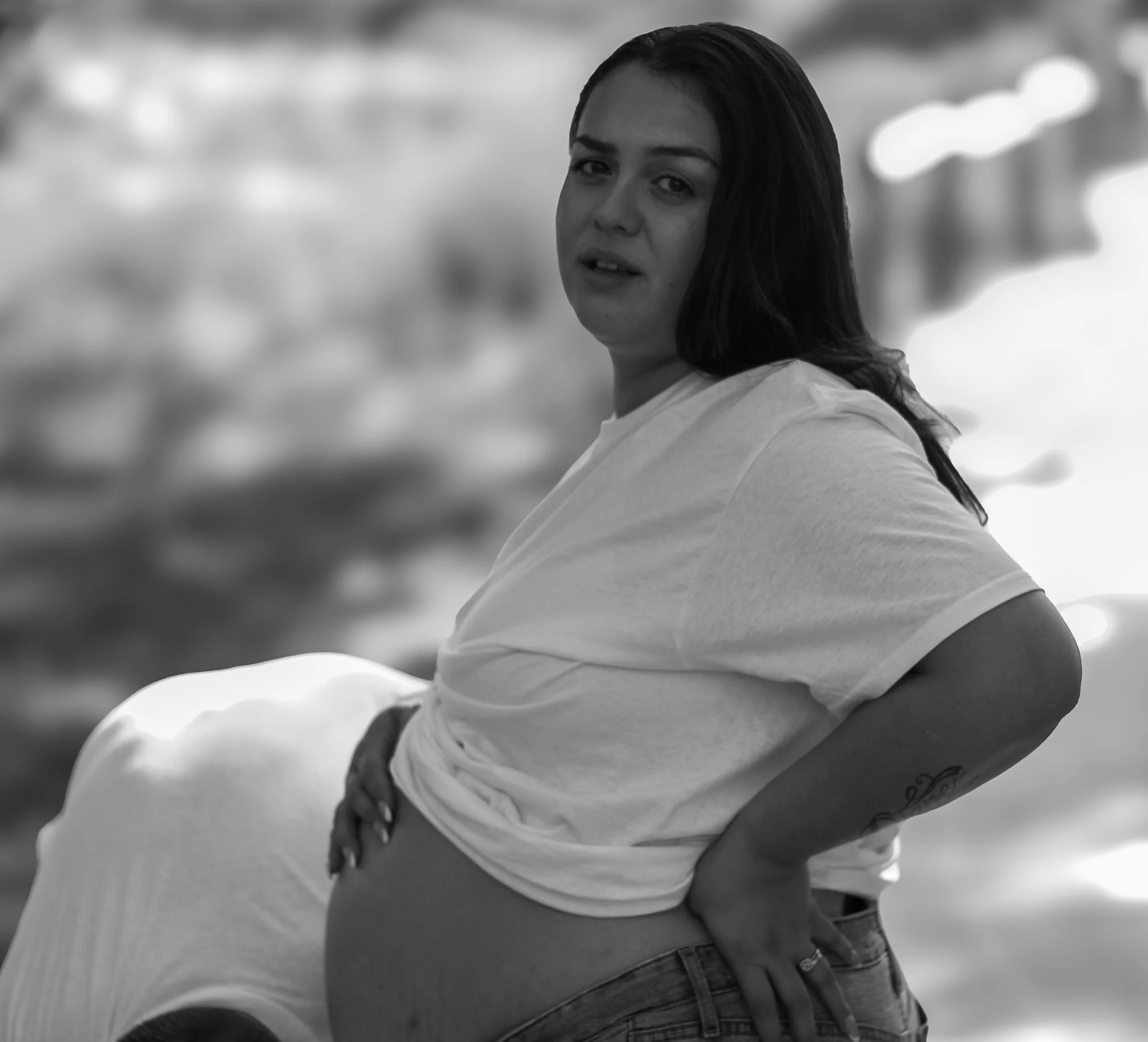 A young woman with wet, dark hair and tattoos on her arm, wearing a white T-shirt and jeans, standing outdoors on a cloudy day.