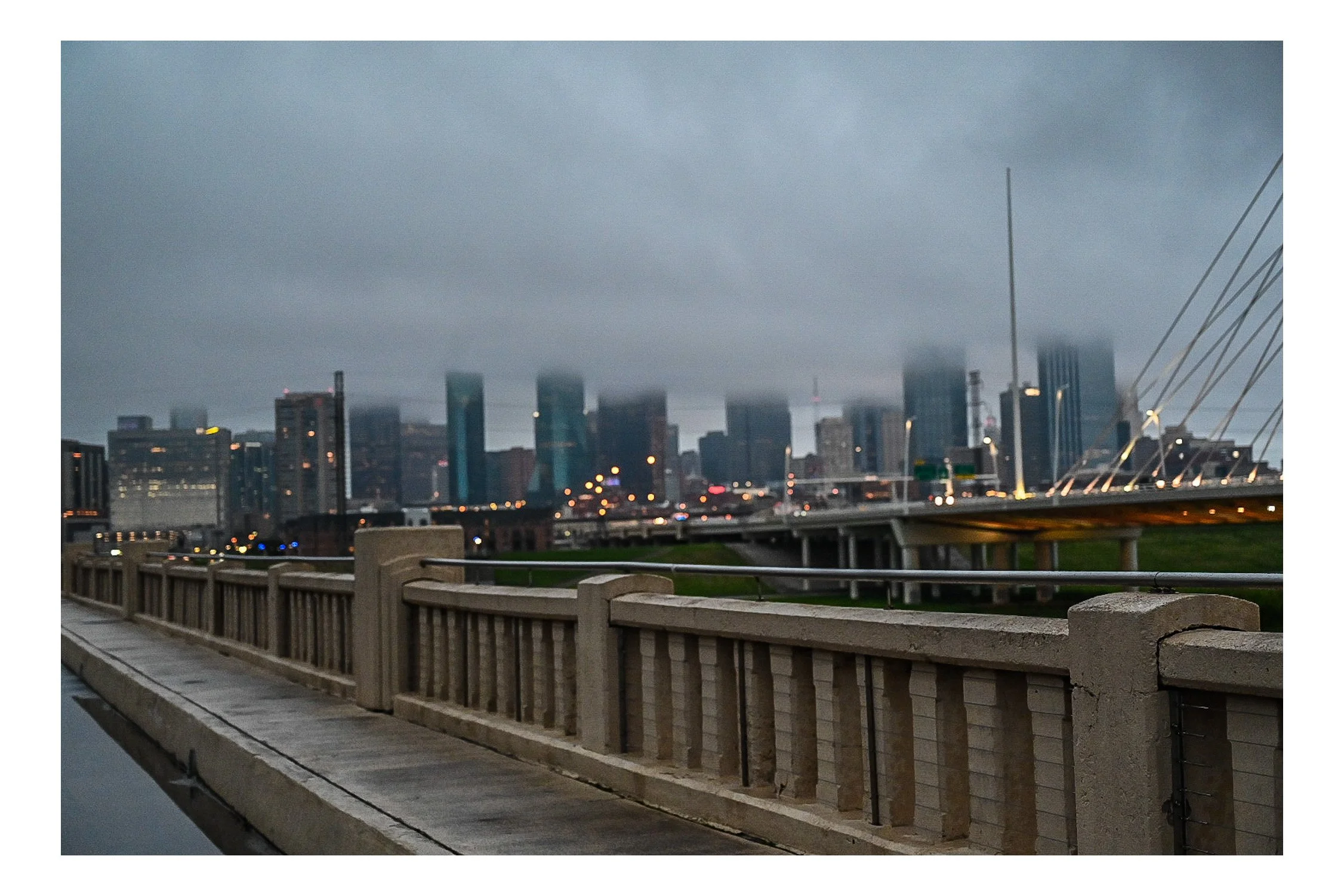 City skyline with tall buildings under cloudy sky, seen from a bridge with a concrete railing and walkway.