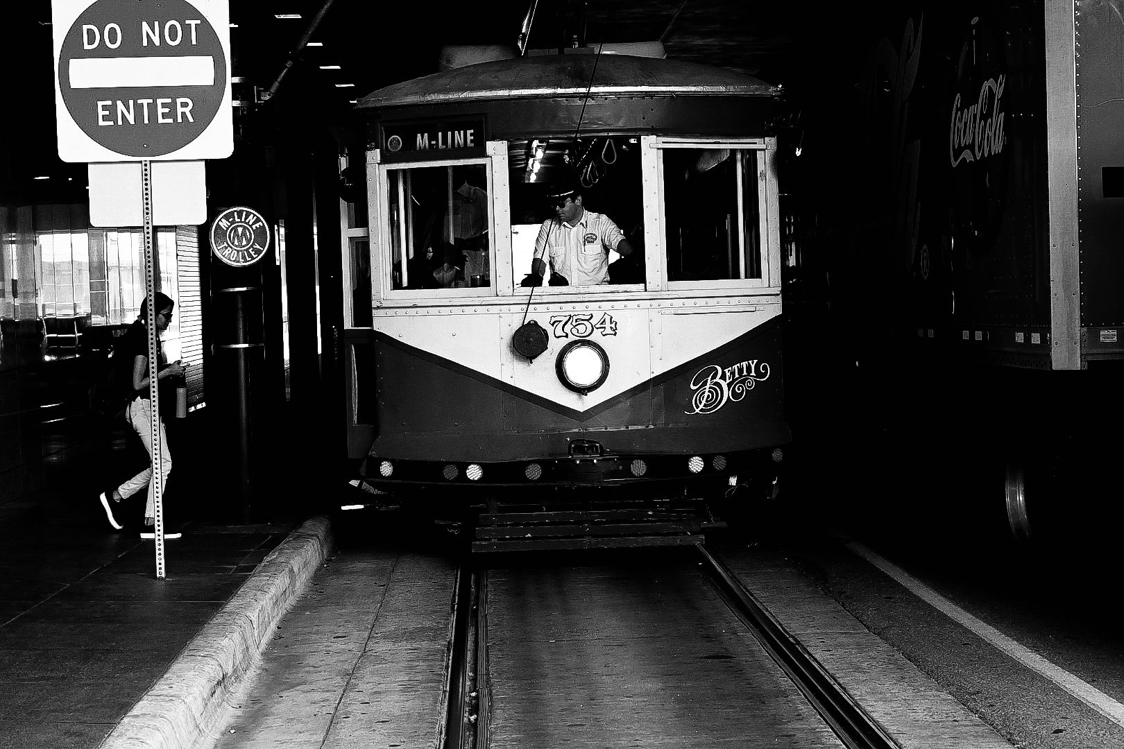 A black and white photo of a vintage trolley on city tracks with a conductor inside. A woman with a backpack is walking on the sidewalk looking at her phone, past a 'Do Not Enter' sign.