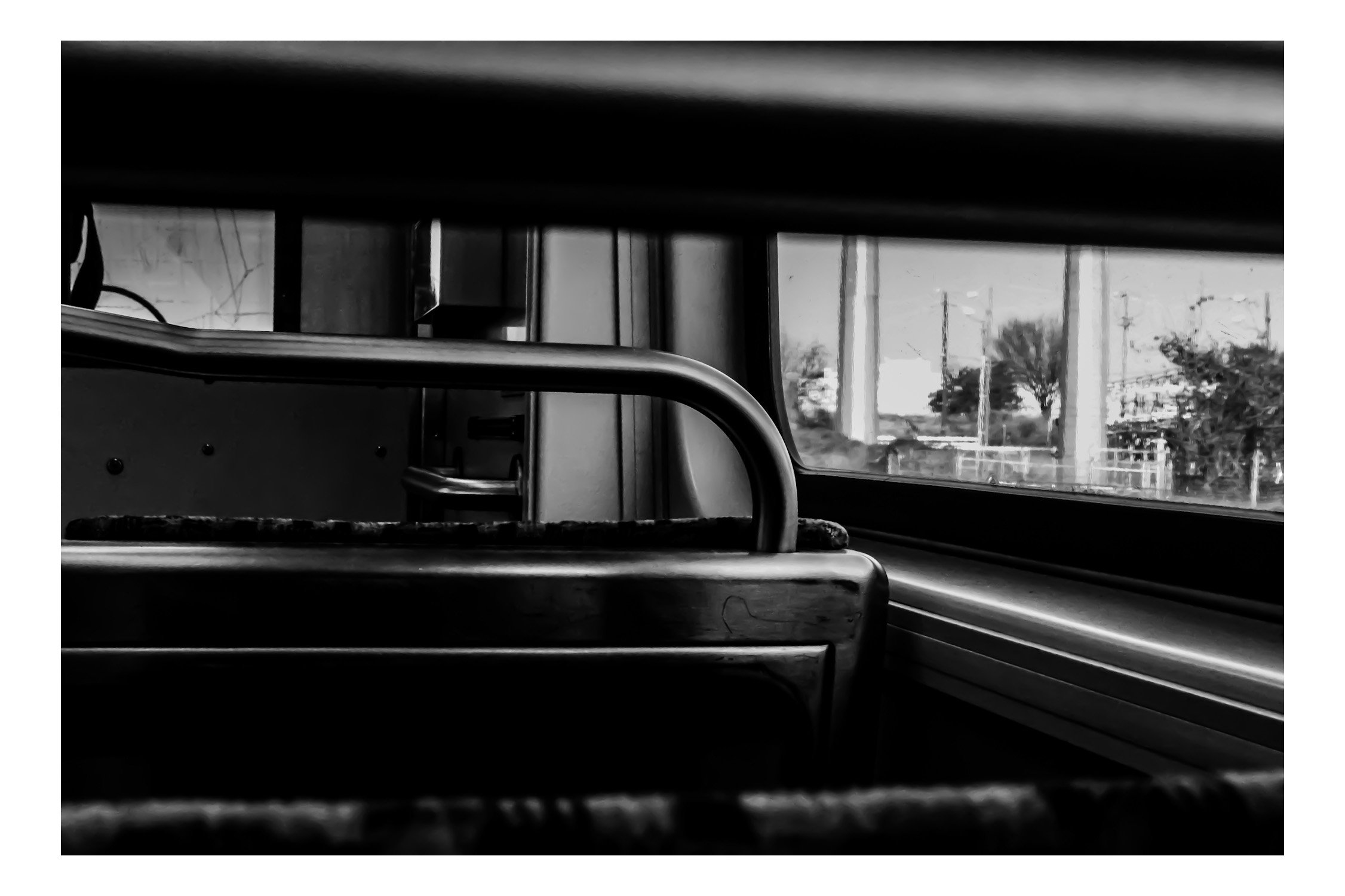 Black and white photo taken from inside a vehicle, looking through a rear window with a metal handle in the foreground. Outside, power lines, trees, and utility poles are visible.
