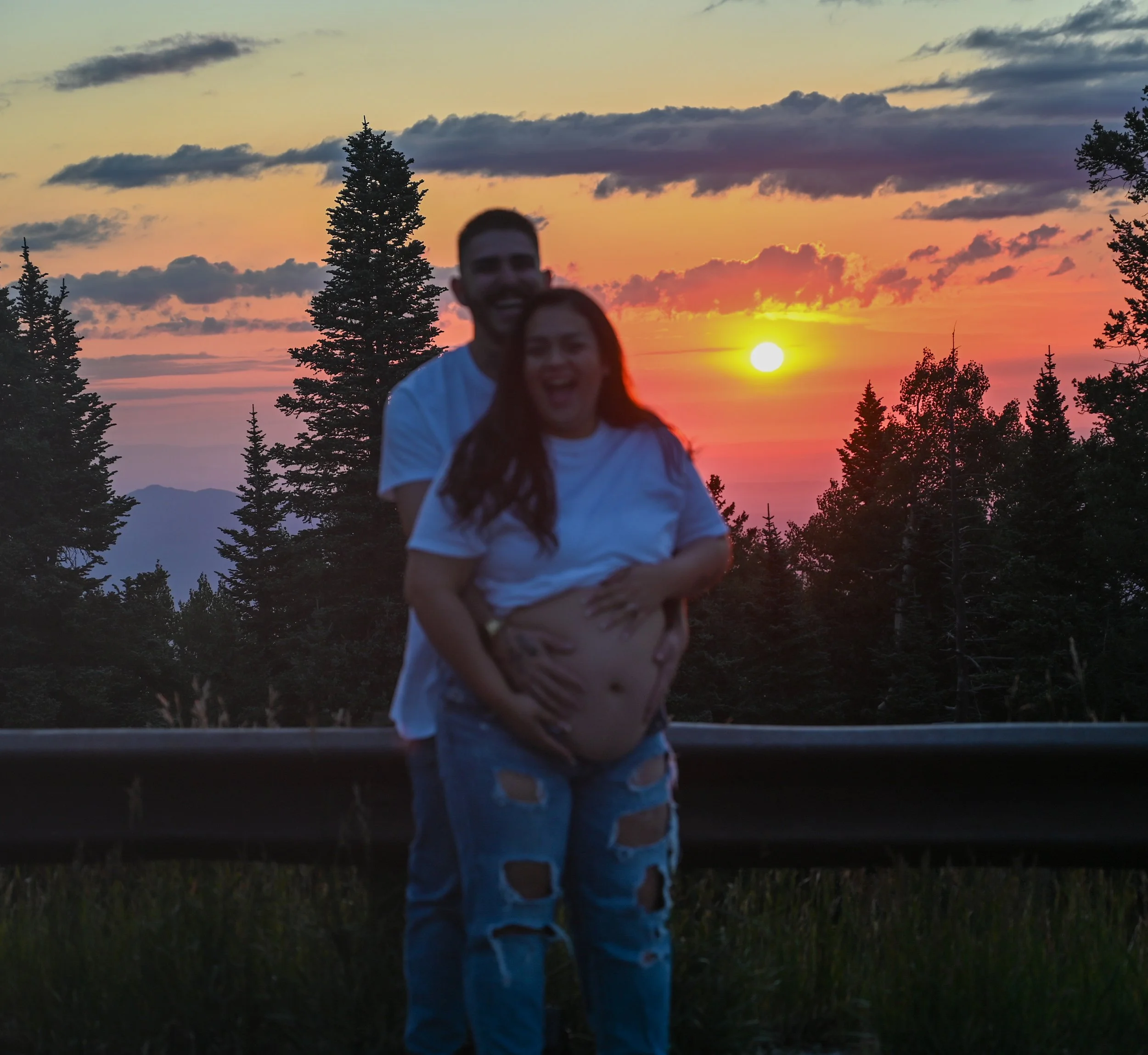 Two people, a man and a woman, standing close together outdoors during sunset. The scene includes tall pine trees and a colorful sky with orange, yellow, and purple hues.