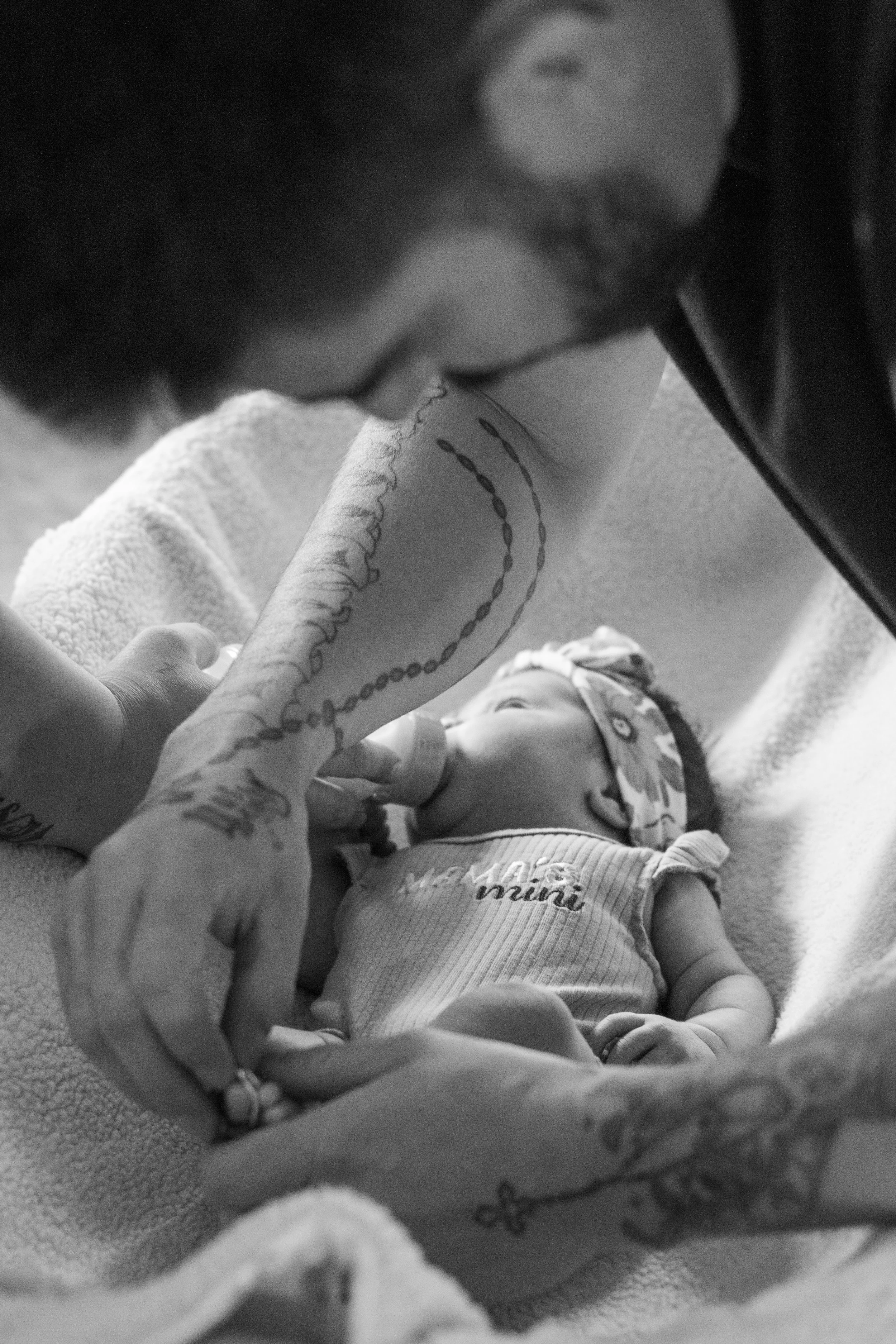 A close-up black and white photo of a person with tattoos on their arm, holding the hand of a baby, who is lying on a soft blanket and wearing a bib and clothing with text. The person is leaning over the baby, and the baby's face is partially visible