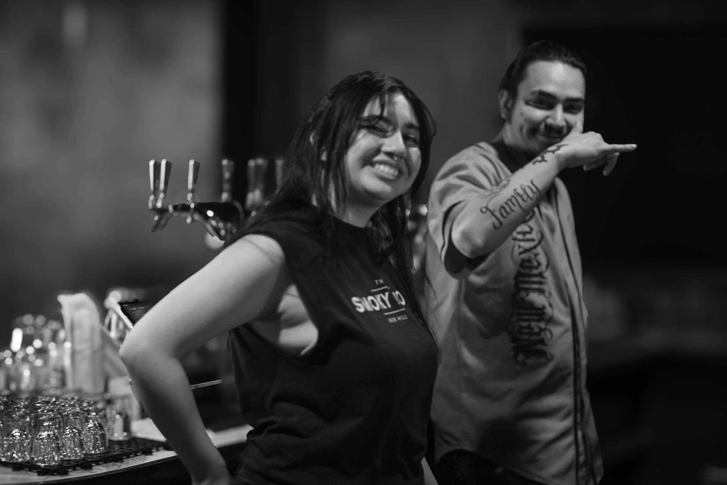 Two happy people, a woman and a man, standing behind a bar counter with glasses and bottles, smiling at the camera in a black and white photo.