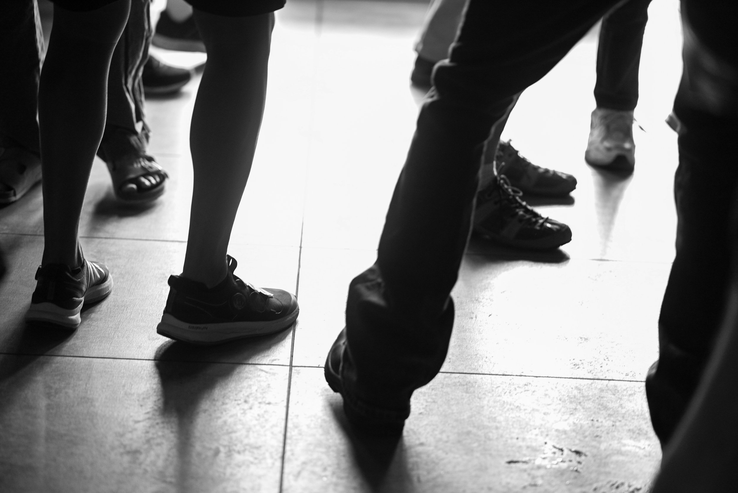 Black and white photo of people's legs and feet, some wearing sneakers, standing on a tiled floor.