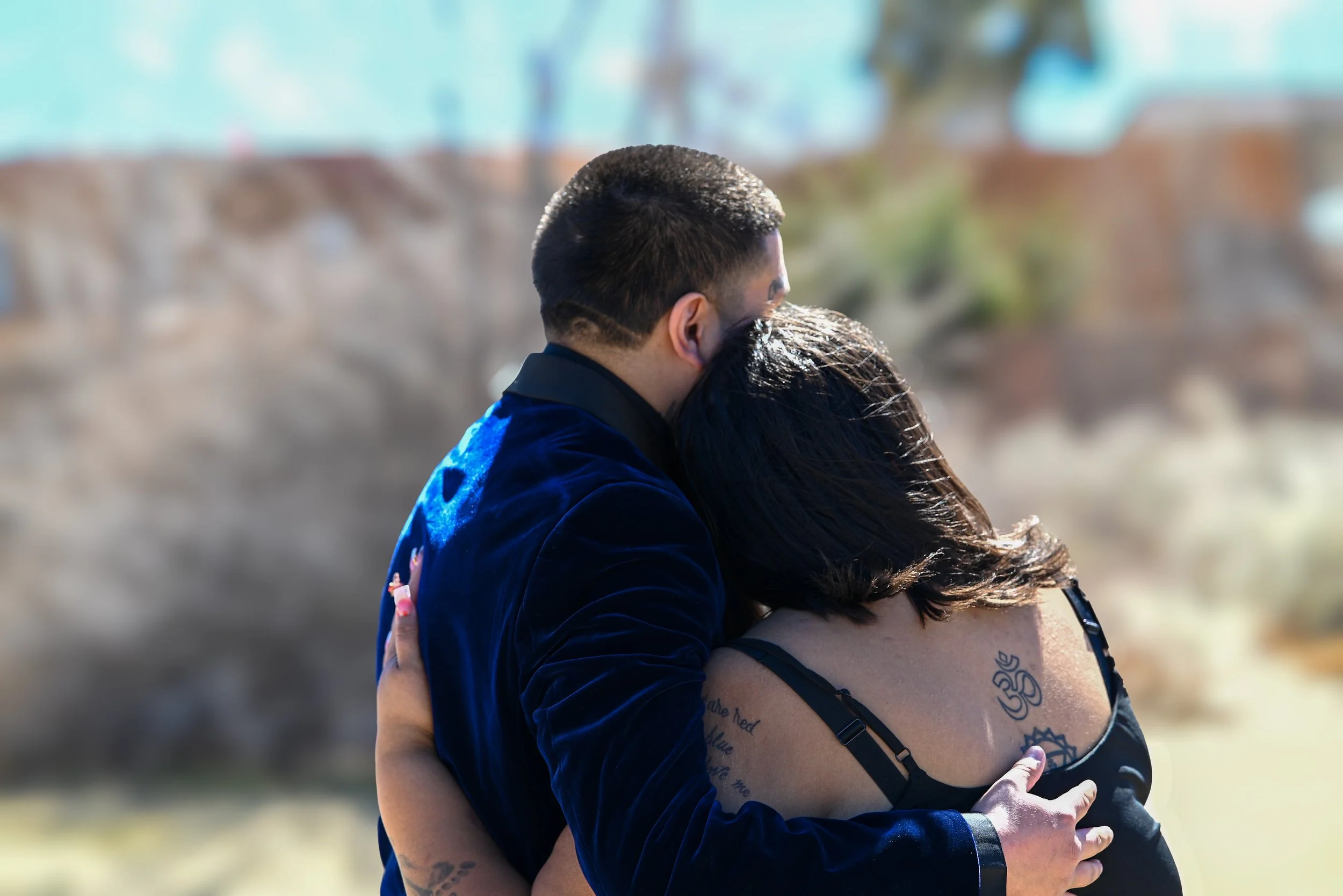 A couple is embracing outdoors, with the man wearing a dark jacket and the woman in a black top, showcasing tattoos on her arm and shoulder.