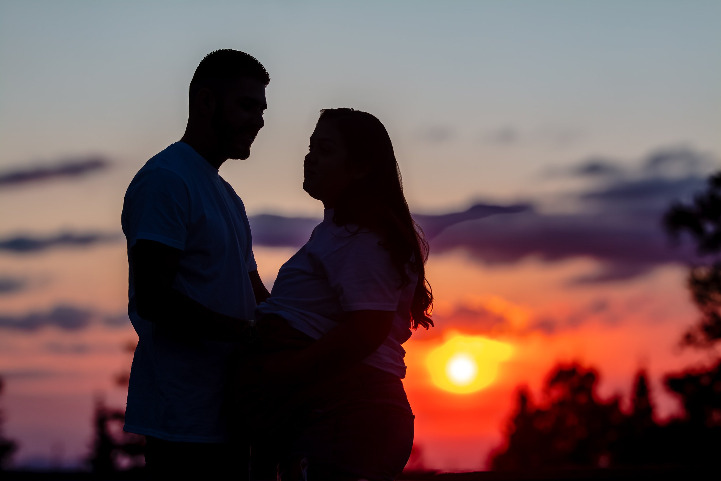 A silhouette of a man and woman standing close together outdoors at sunset, with colorful sky and blurred landscape in the background.