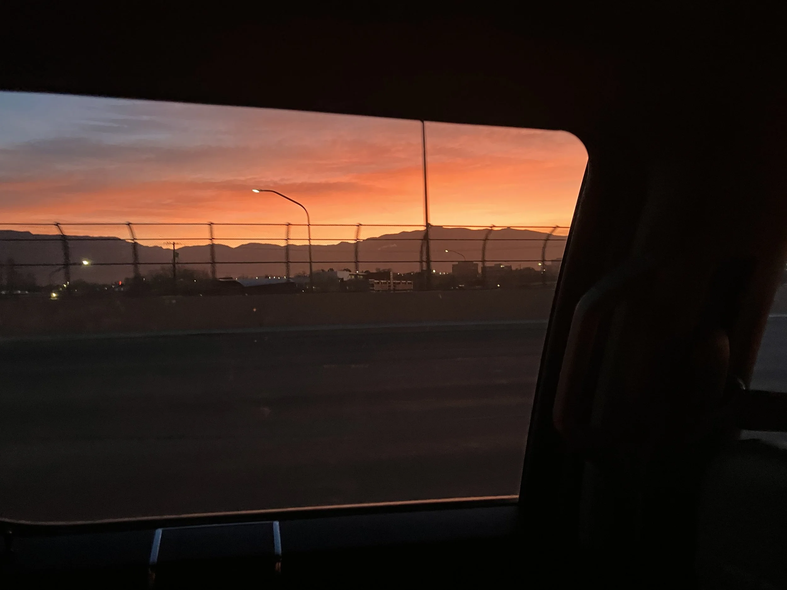 View of a sunset sky with orange and pink hues seen through the window of a moving vehicle, with a fence and mountains in the distance.