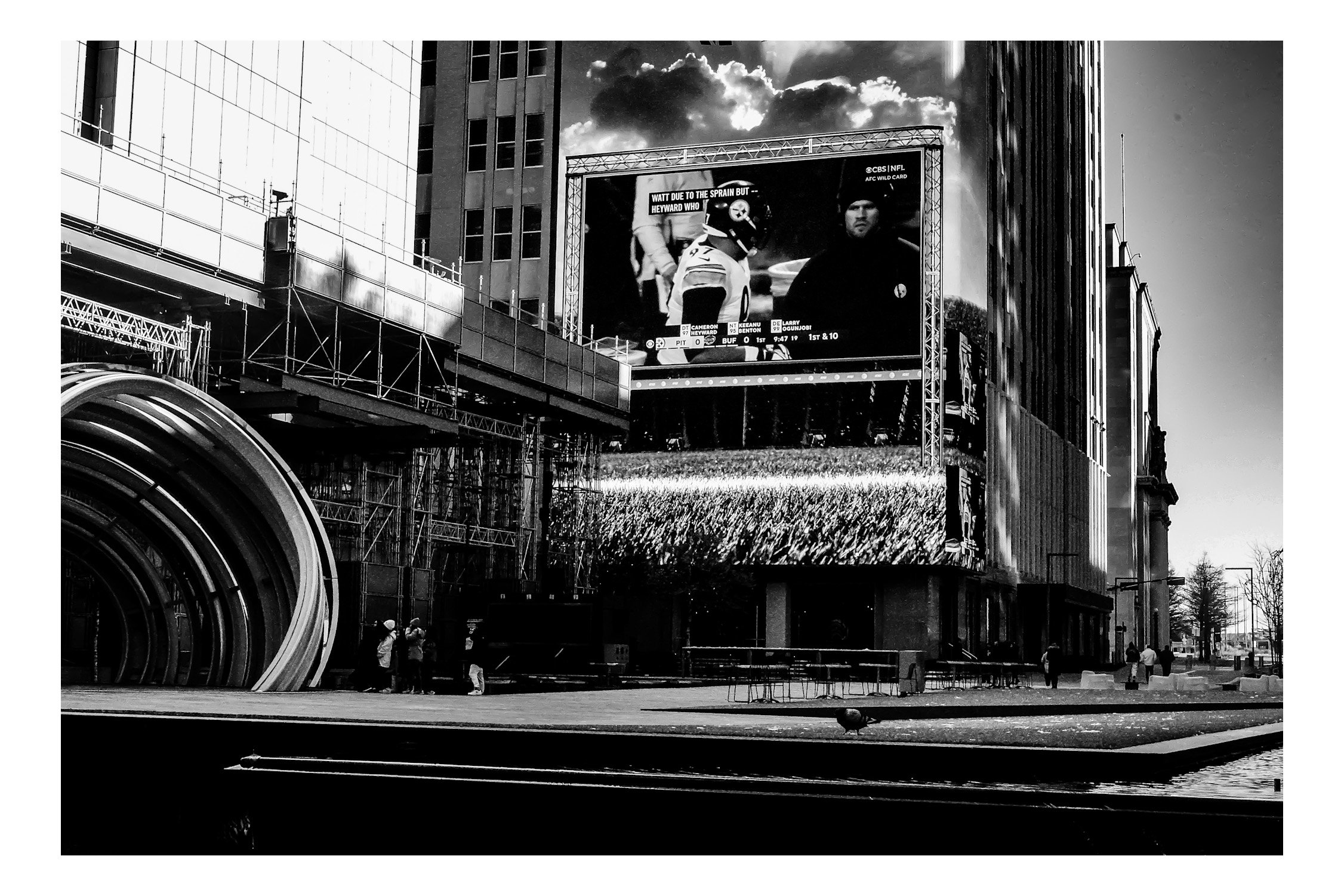 A black and white cityscape with tall buildings, a large digital billboard displaying a football game, construction scaffolding, and people walking on the street.