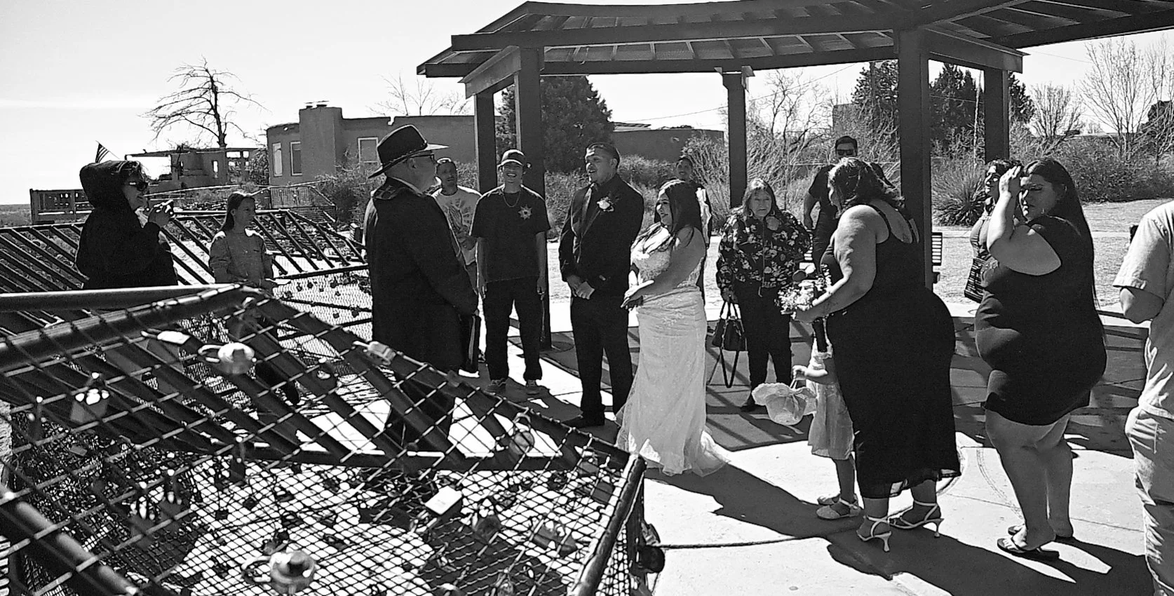 Group of people gathered outdoors under a shelter, with a woman in a white dress and a man in a suit at the center, possibly during a wedding ceremony.