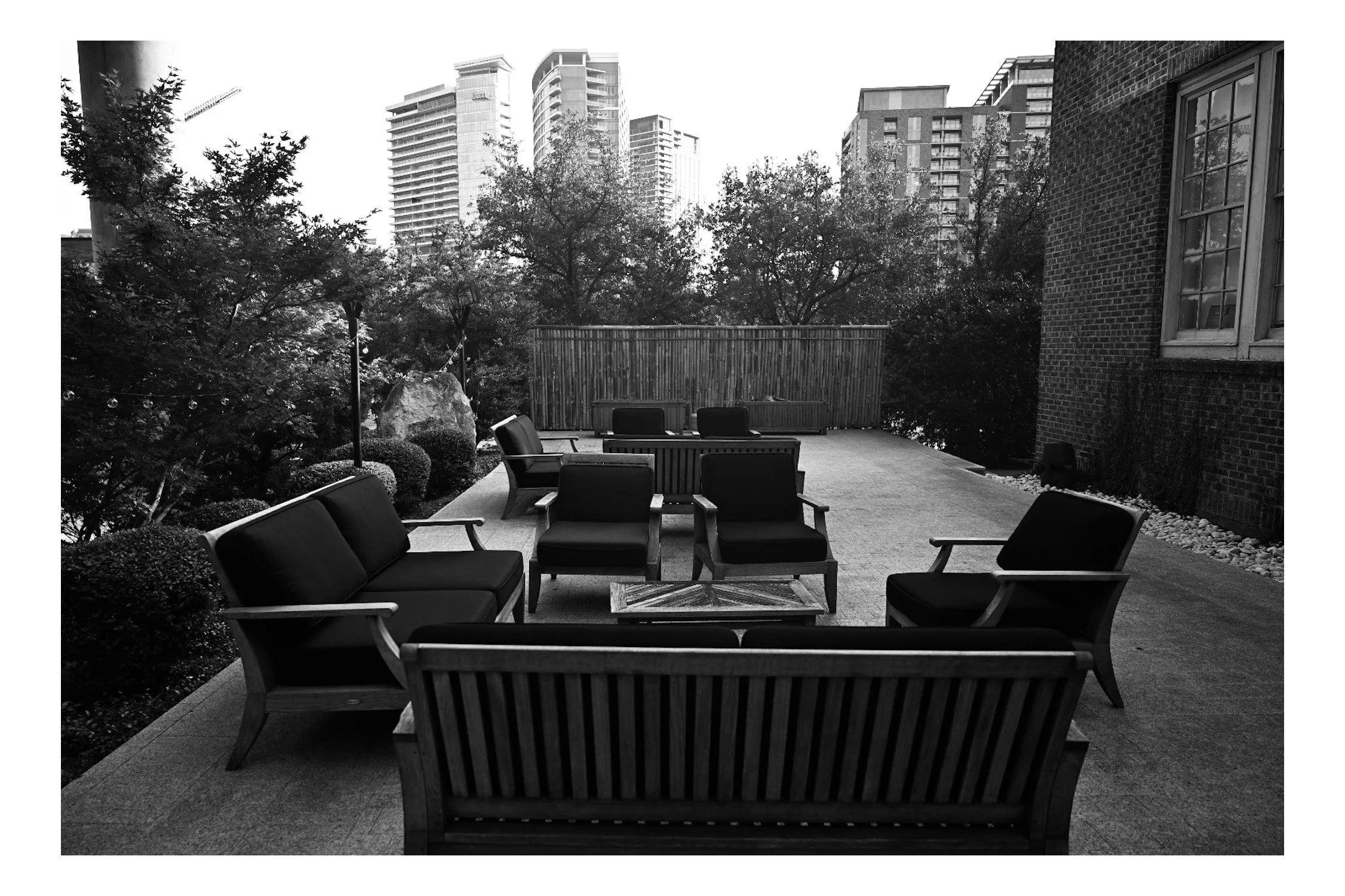 An outdoor balcony with multiple chairs surrounding a low table, with trees and high-rise buildings in the background.