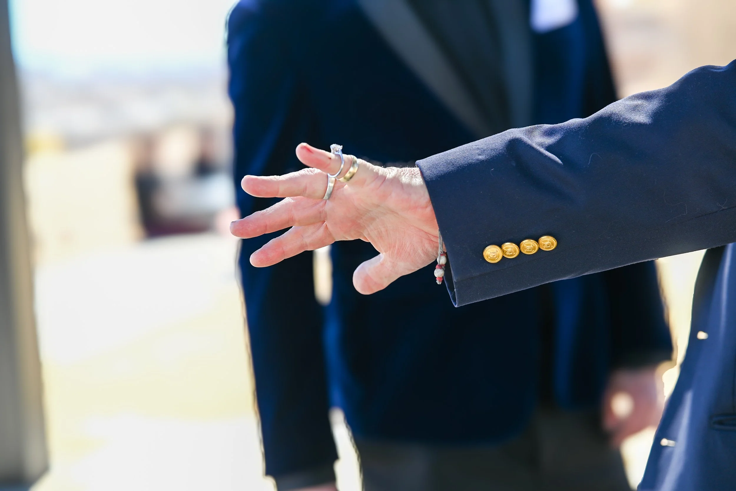 Close-up of two people shaking hands, one wearing a navy blazer with gold buttons, the other wearing a dark suit and jewelry, in an outdoor setting with bright natural light.