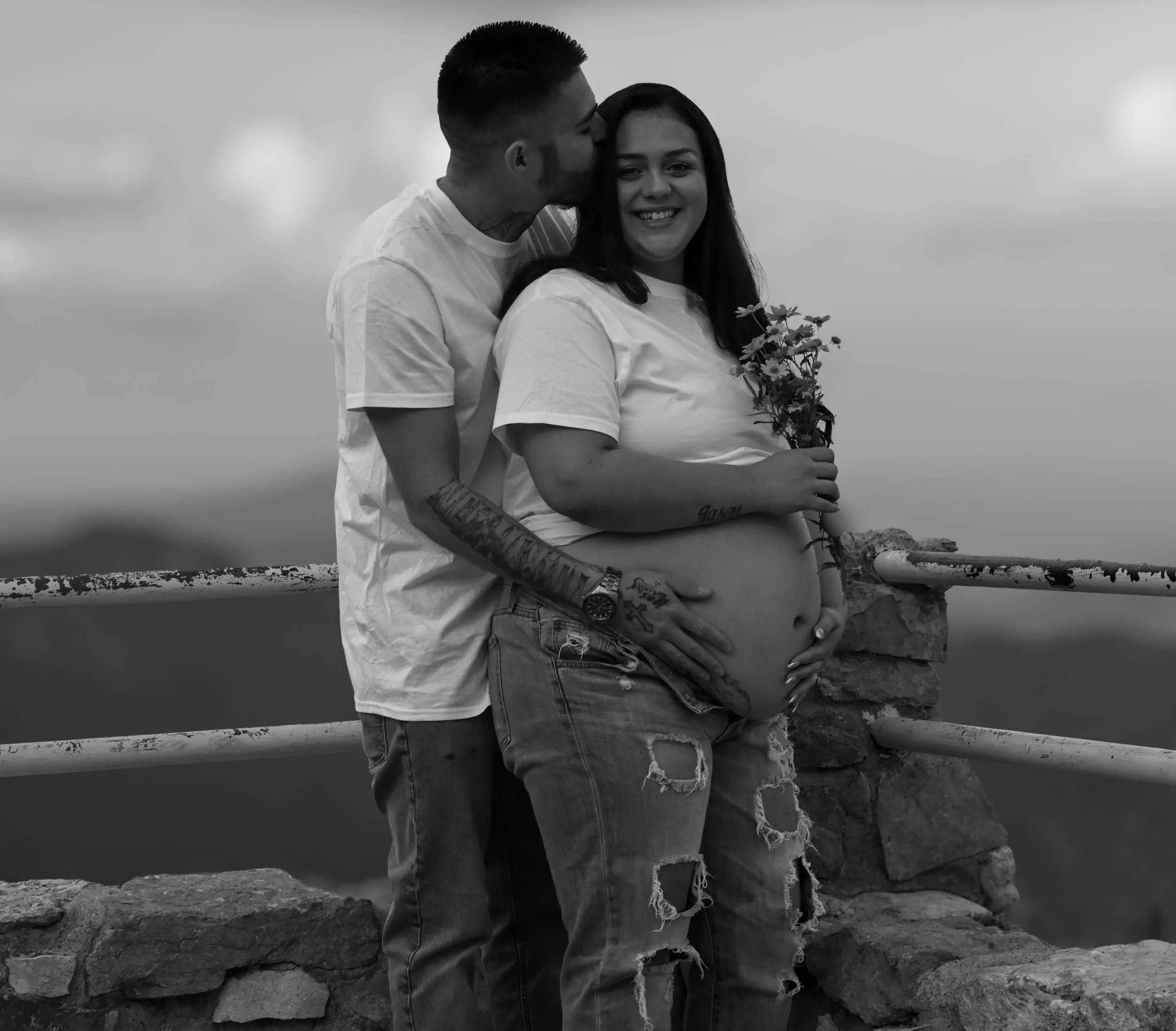 A couple stands close together outdoors, with the man kissing the woman's forehead. She holds a small bouquet of flowers and smiles at the camera. The photo is in black and white.