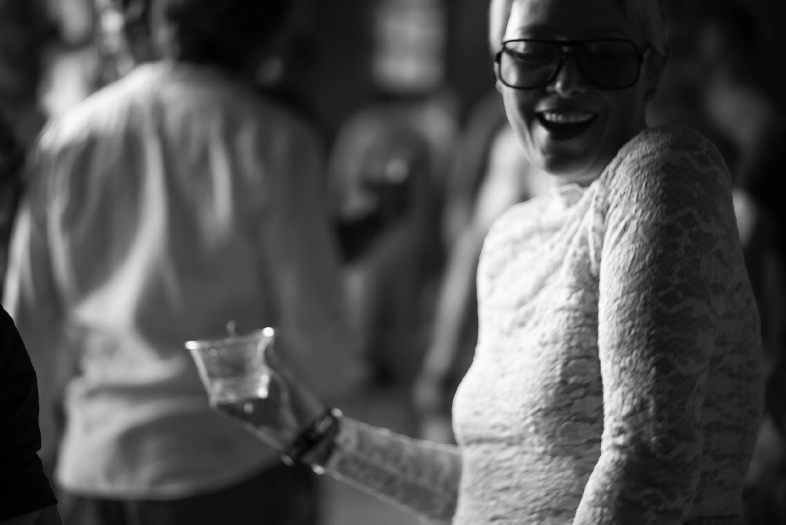 A woman with short blonde hair, wearing glasses and a white textured sweater, smiling and looking to her right. The background shows blurred figures, possibly at a social gathering or event, in black and white.