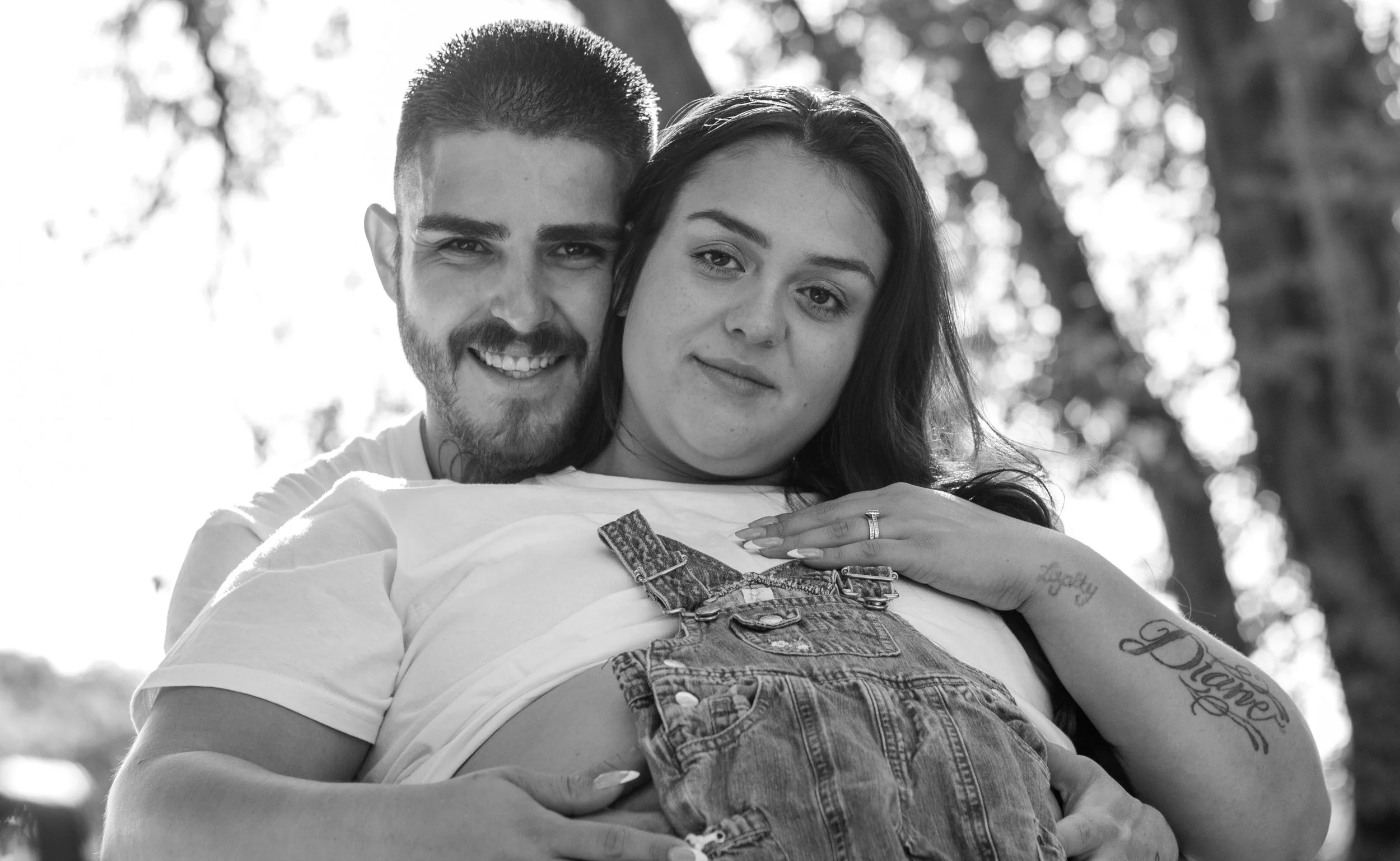 A black and white photo of a couple embracing outdoors, with trees in the background. The man is smiling and the woman has a relaxed expression, with her arms around the man's shoulders.