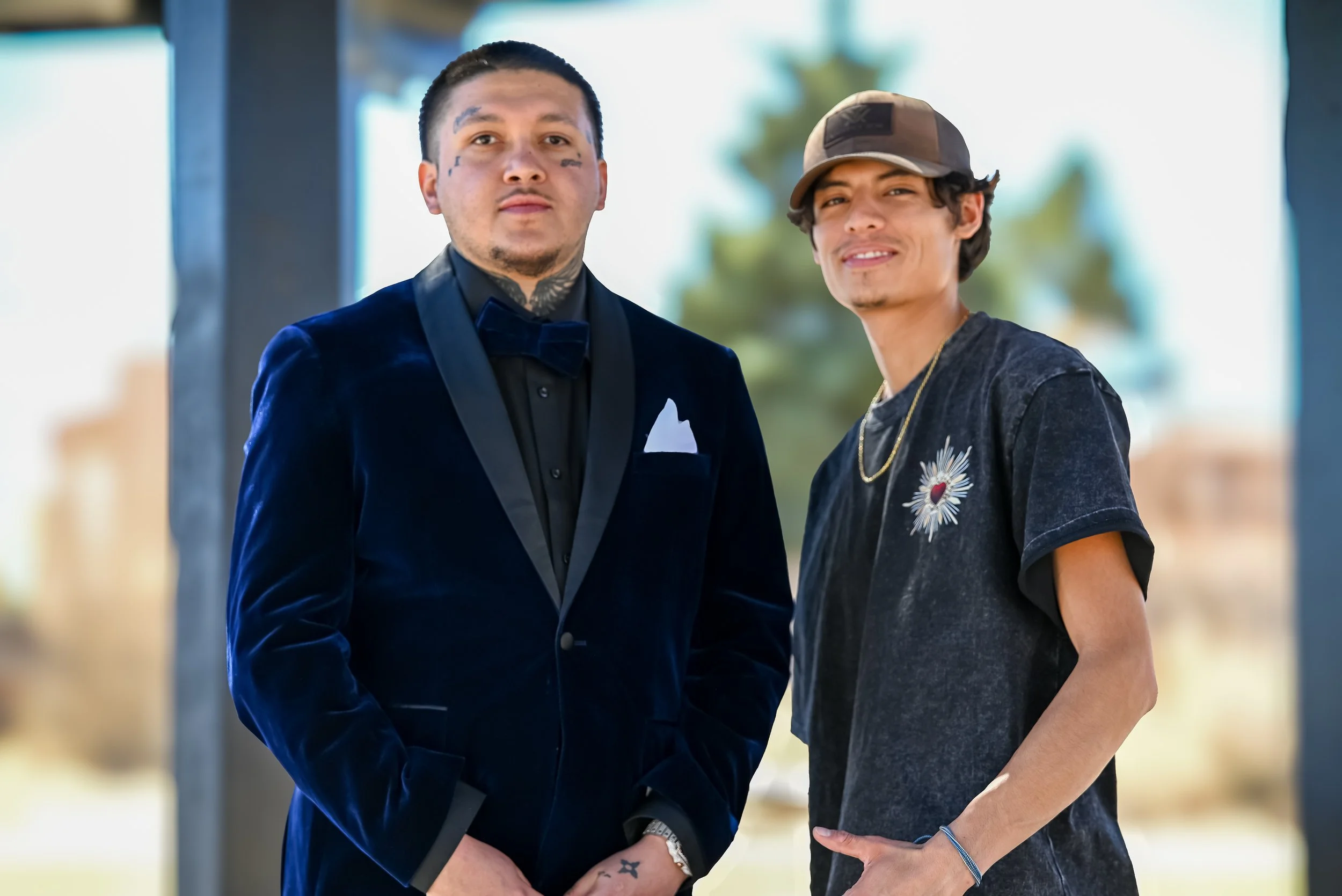 Two men standing side by side outdoors. The man on the left is wearing a dark blue velvet tuxedo with a black shirt and bow tie, and has tattoos on his face and neck. The man on the right is dressed casually in a black T-shirt with a floral design, a