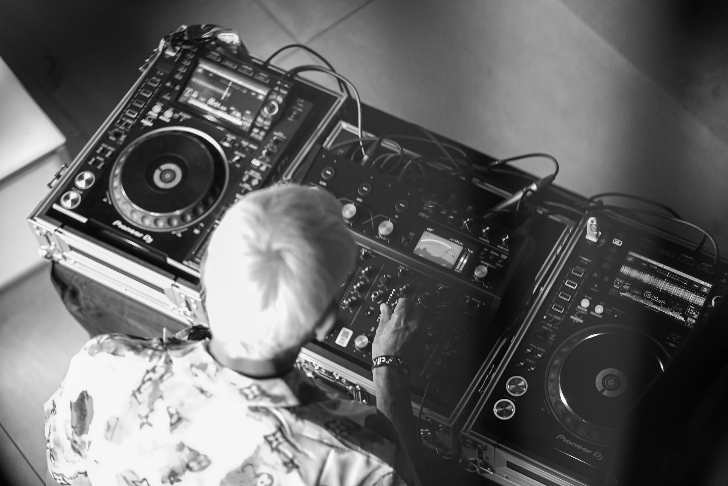A person with light-colored hair operating DJ equipment, including a Pioneer DJ controller, in a black-and-white photo.