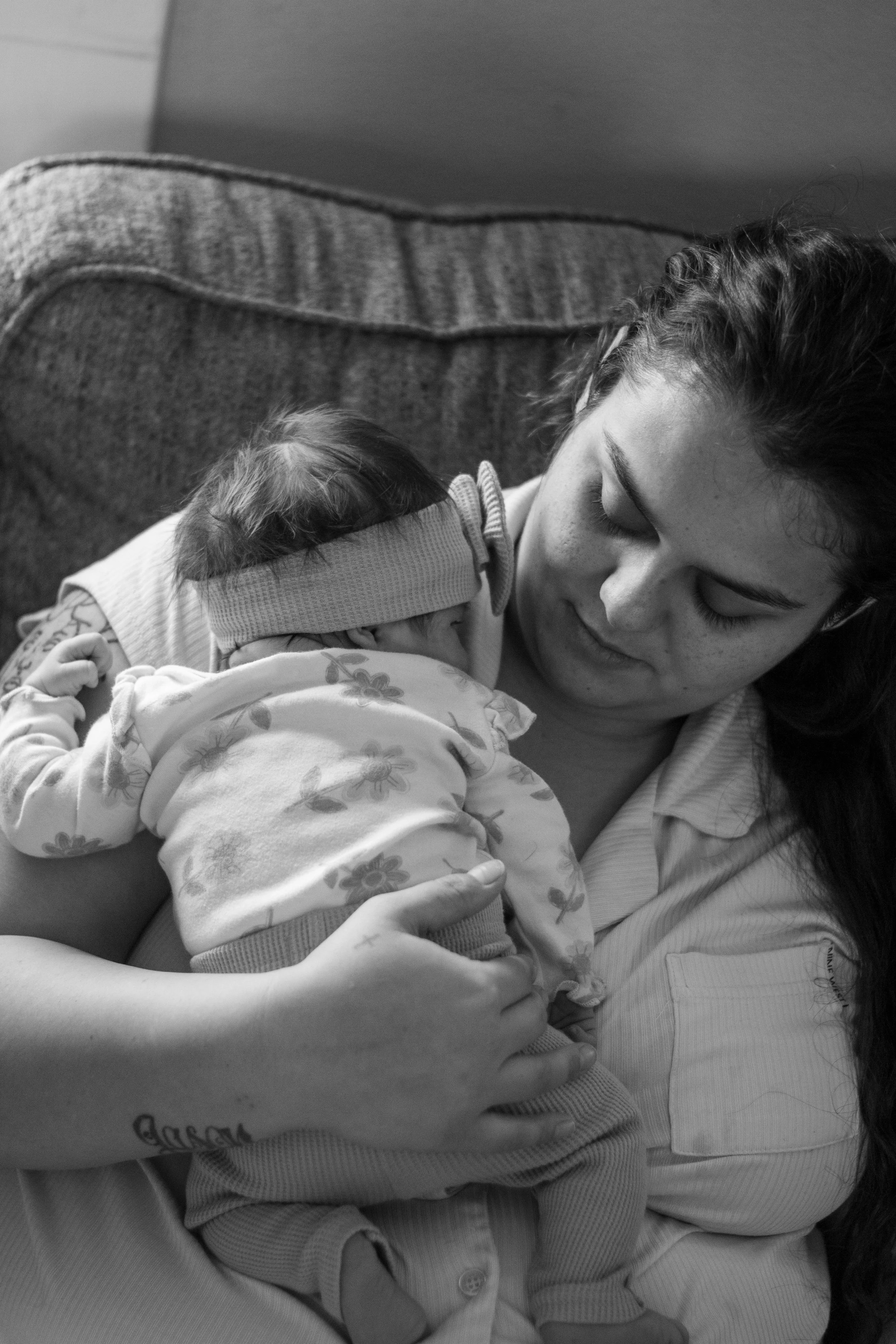A woman with curly hair holding a young child, likely a girl, while lying on a couch. The woman is looking at the child, who is wearing a floral-patterned outfit. The photo is in black and white.