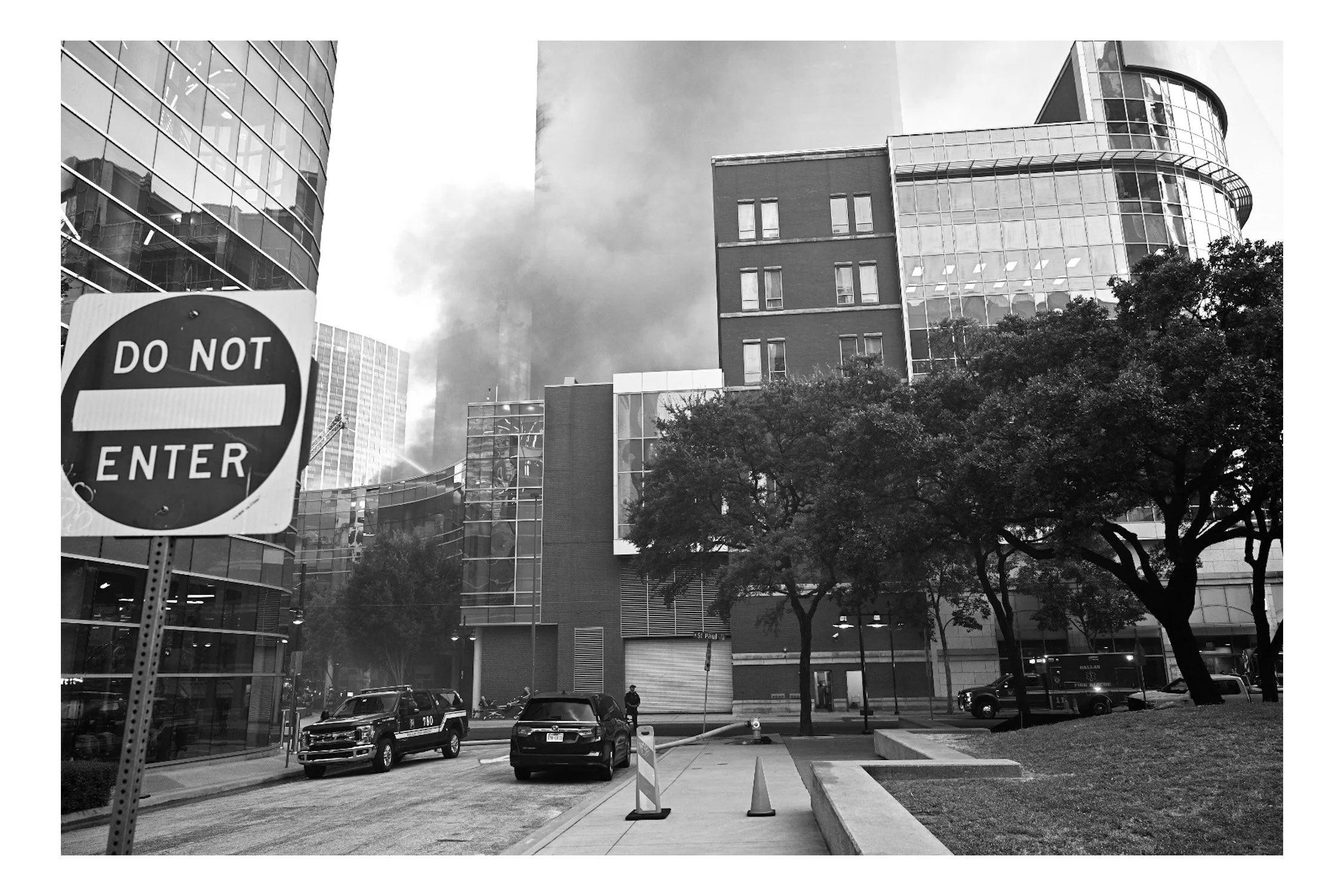 Cityscape with modern glass buildings, a parking lot with cars, a DO NOT ENTER sign, and trees in the foreground. Overcast sky.