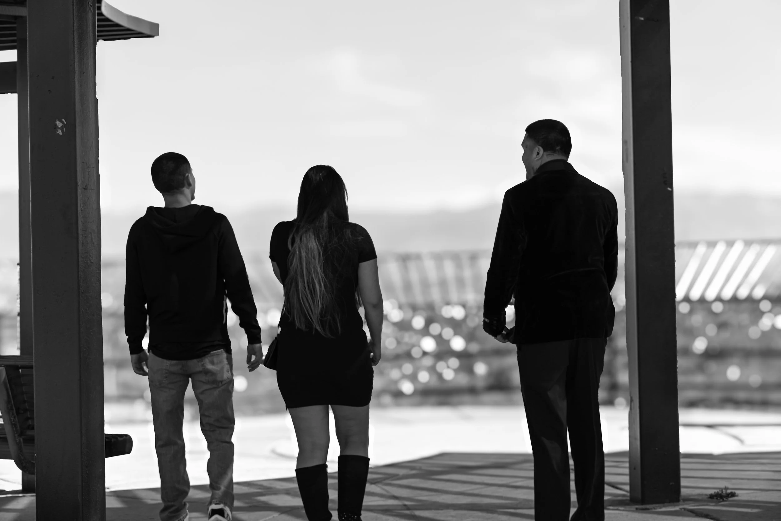 Four people, two men and two women, are standing together on a balcony overlooking the city at dusk, framed by vertical beams. The scene is in black and white.
