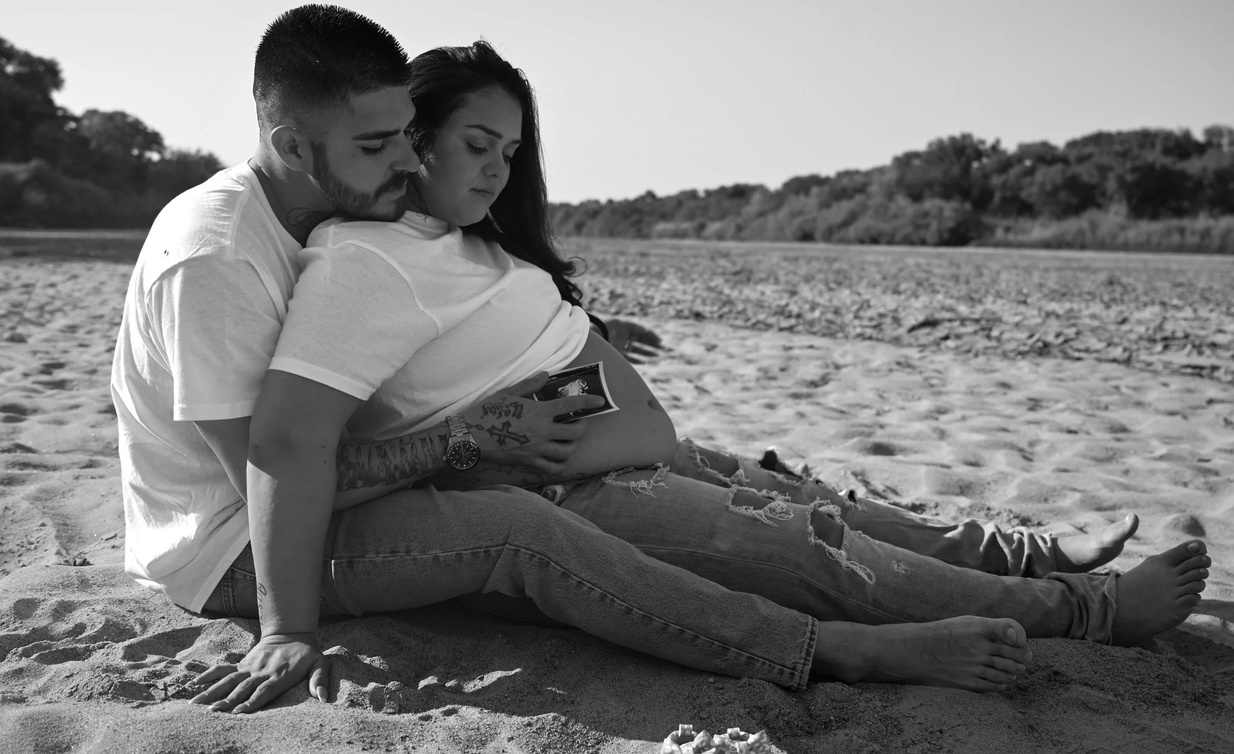 A black and white photo of a man and woman sitting on the beach, close together, looking at a photo or phone. The man is behind the woman, with his arms around her, and they both look focused. The background shows a sandy beach and trees.