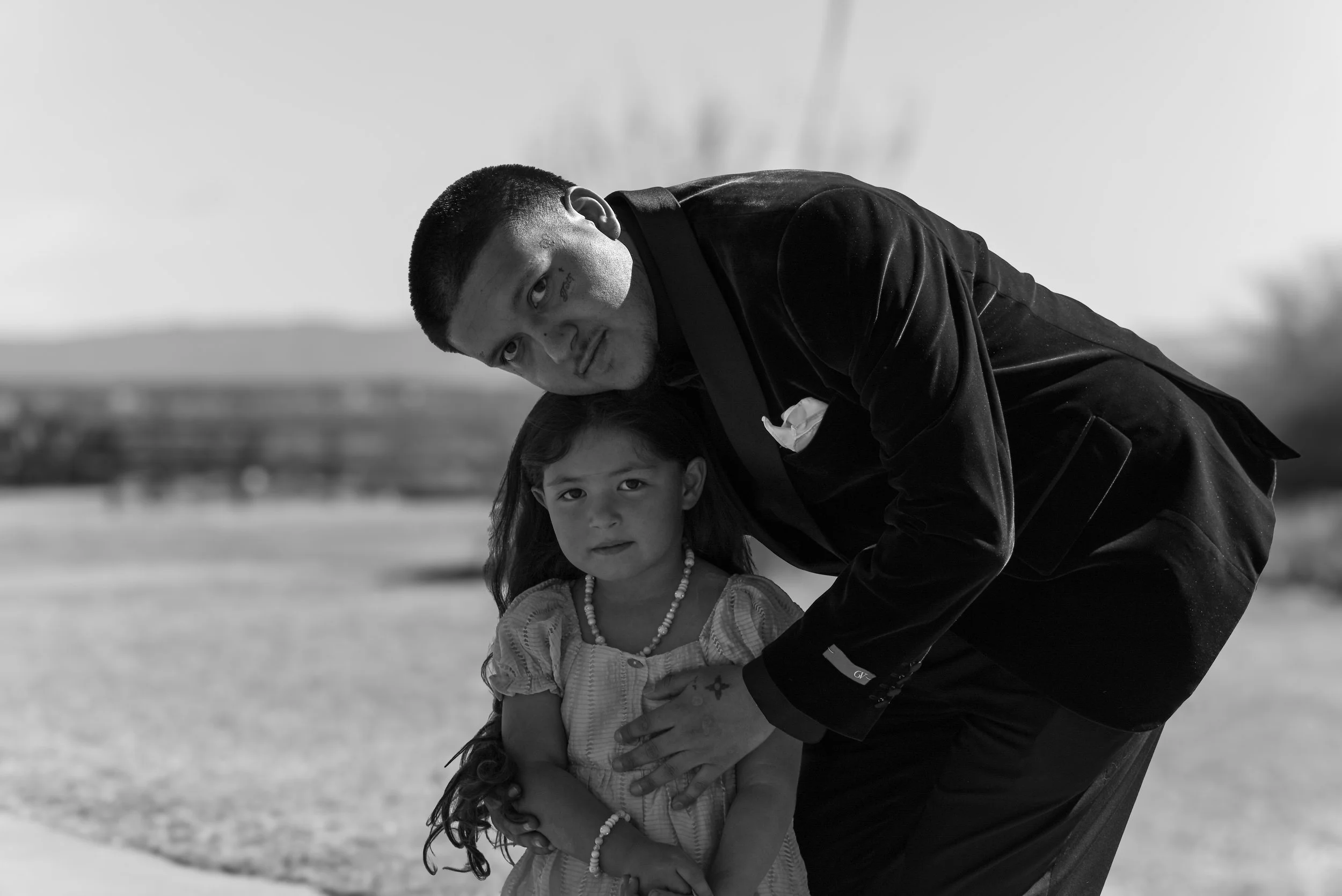 A man and a young girl outside, with the man bending over and looking at the camera while hugging the girl. The girl has long dark hair and is wearing a necklace and a light-colored top. The man has short hair, a tattoo on his face, and is dressed in