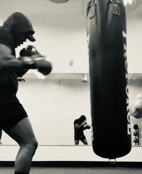 A person in a hoodie and shorts boxing at a punching bag in a gym, with their reflection visible in the mirror behind.