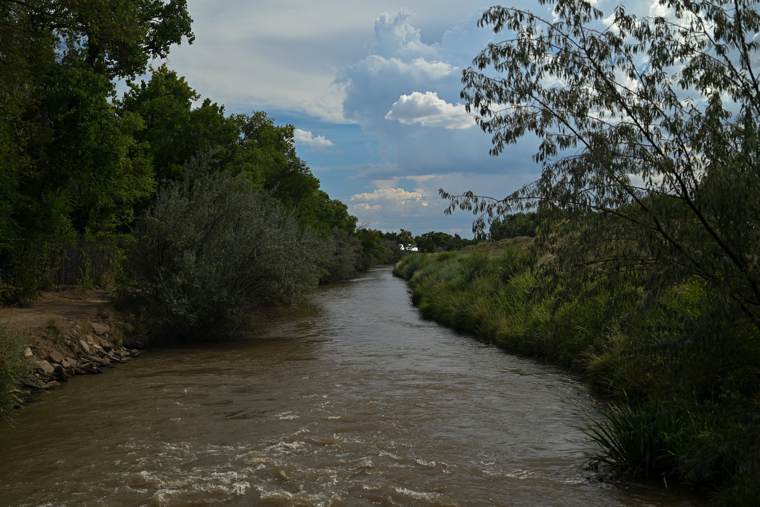 A river flowing through a lush green landscape with trees and bushes on either side, under a partly cloudy sky.