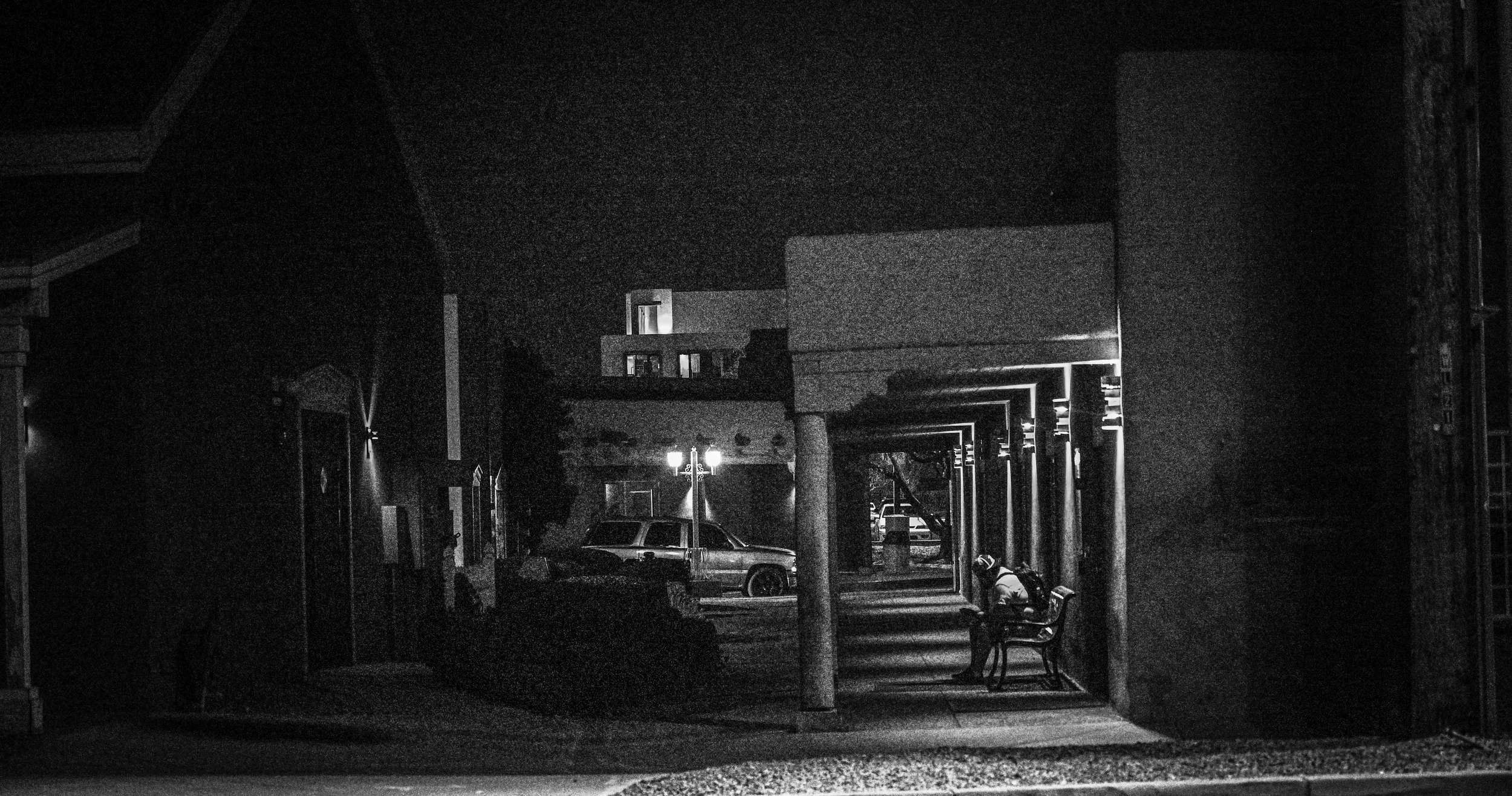 A quiet street scene at night with streetlights, parked cars, and buildings, viewed through an open doorway with benches.