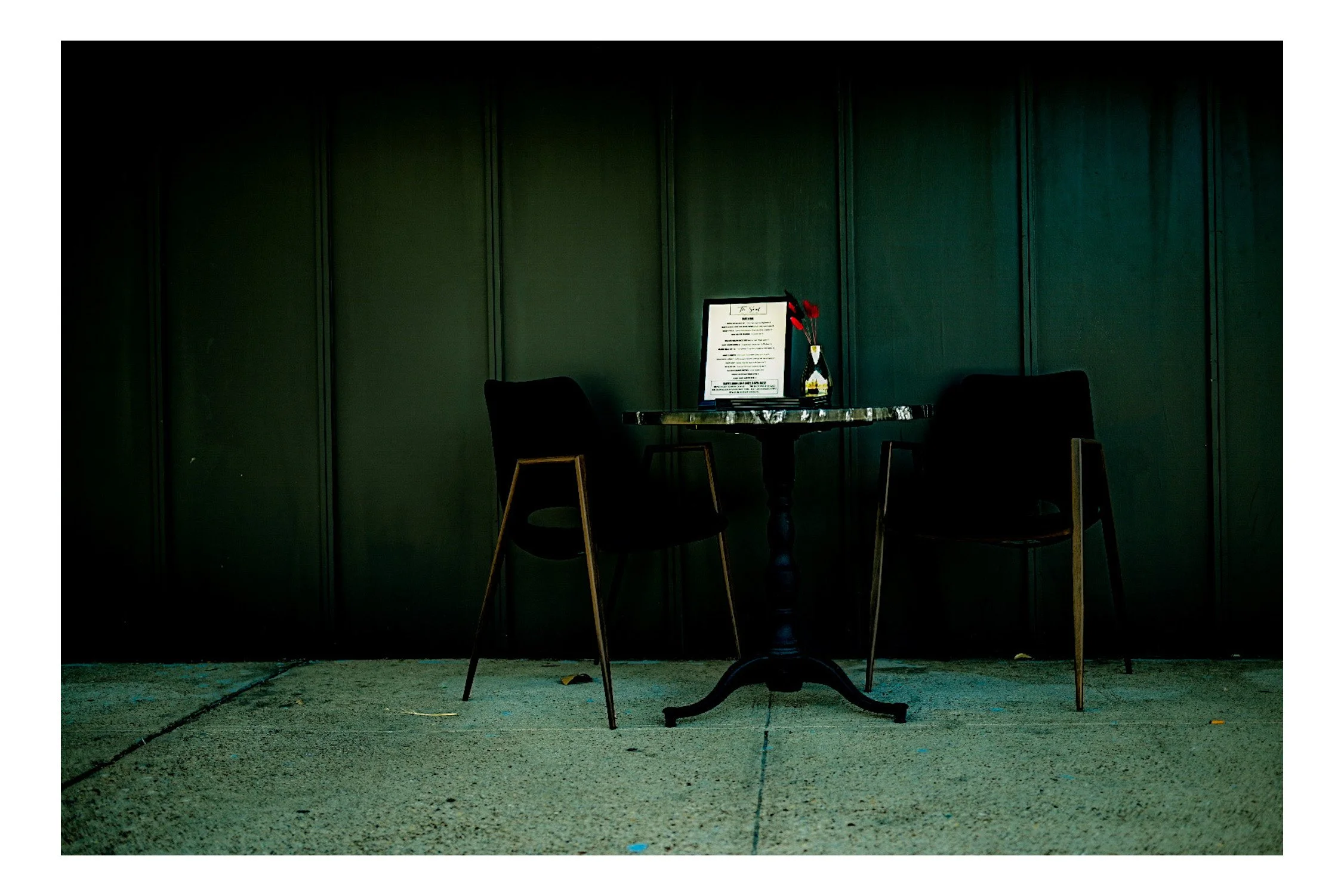 A small round table with a black pedestal base, two black chairs, and a centerpiece with red flowers in a glass vase, set against a dark green paneled wall.