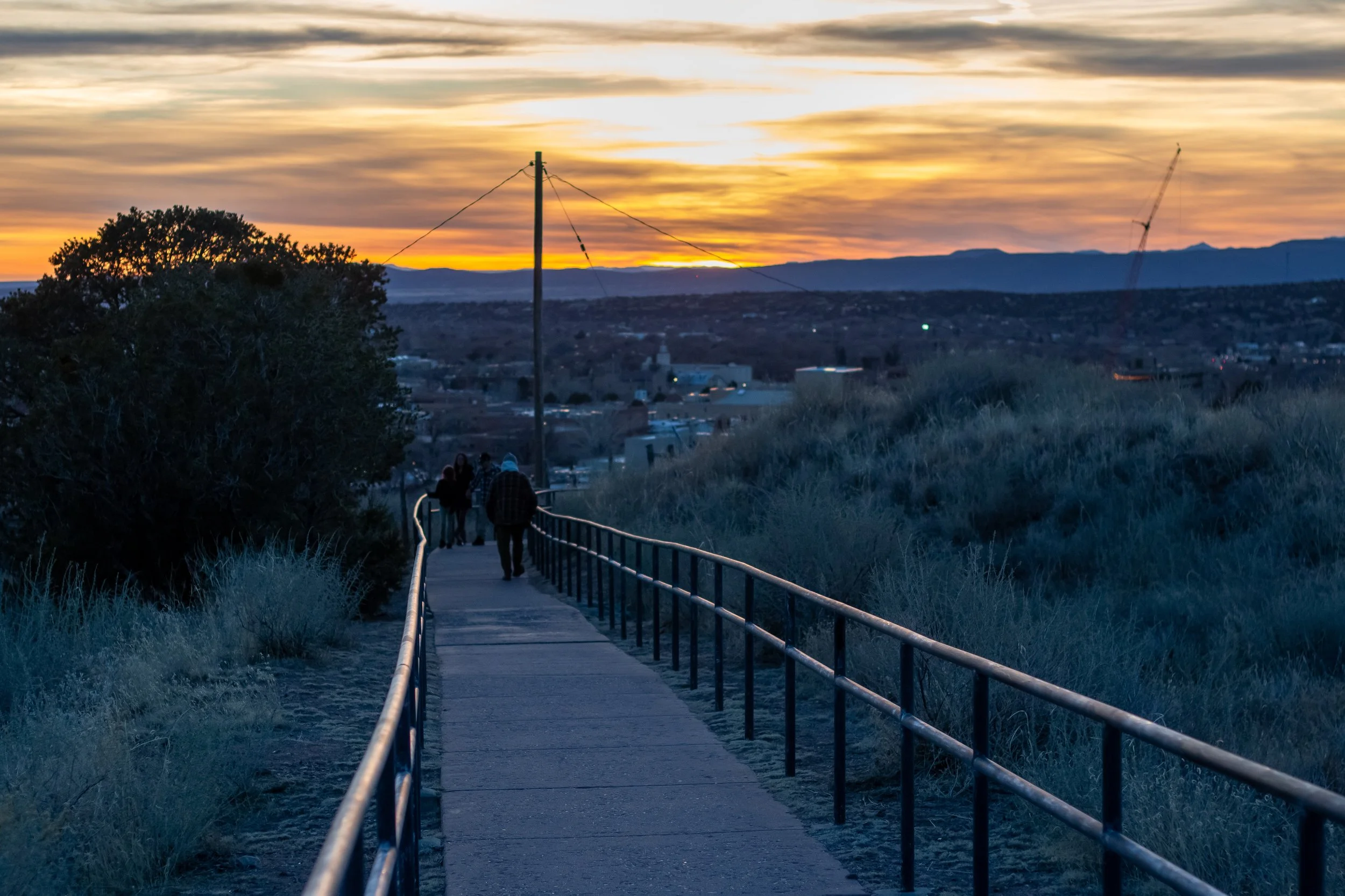A pathway with a handrail during sunset with a cloudy sky, distant mountains, and a few people walking.