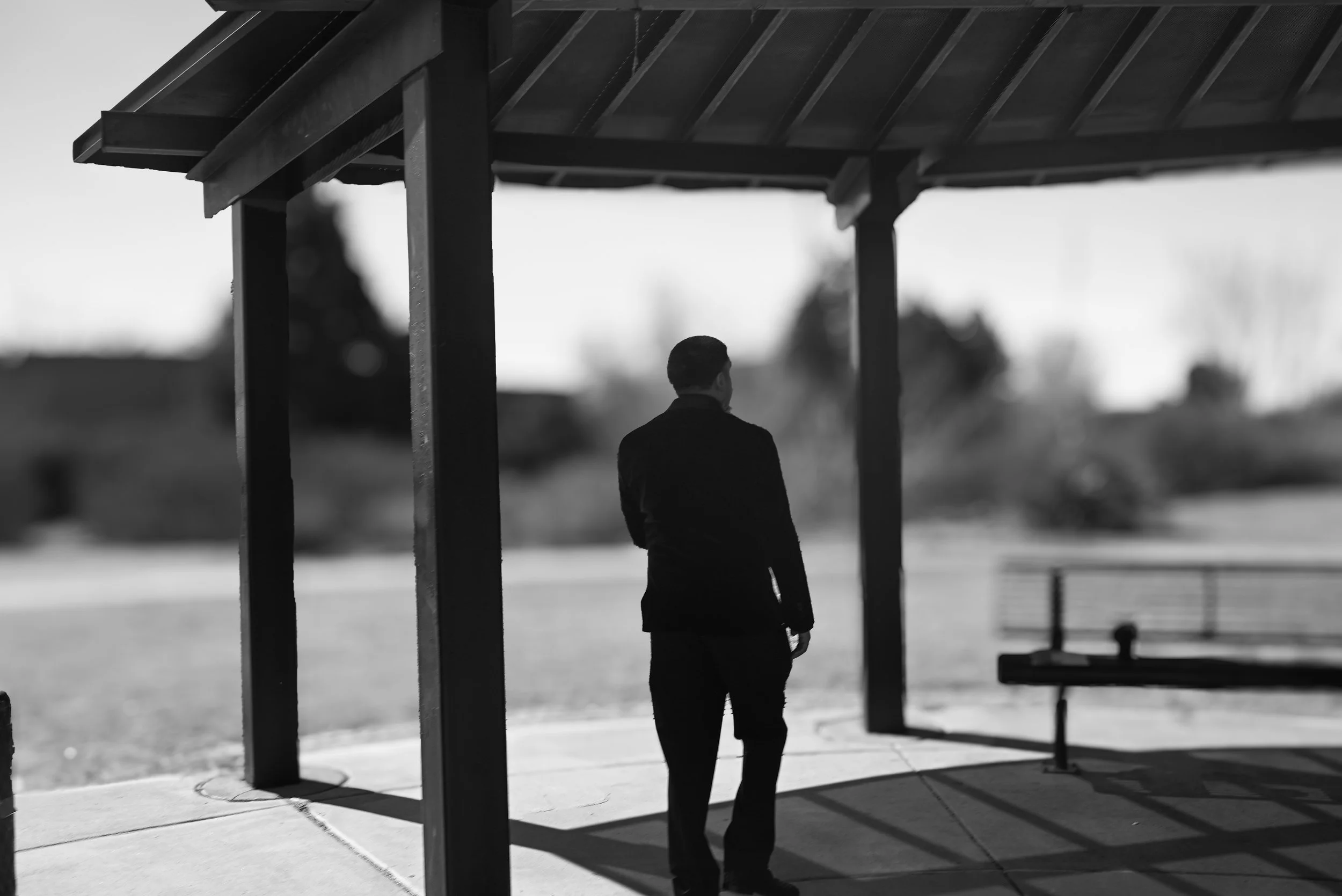 Black and white photograph of a man standing alone under a pavilion at a park, with trees in the background and shadows cast on the ground.