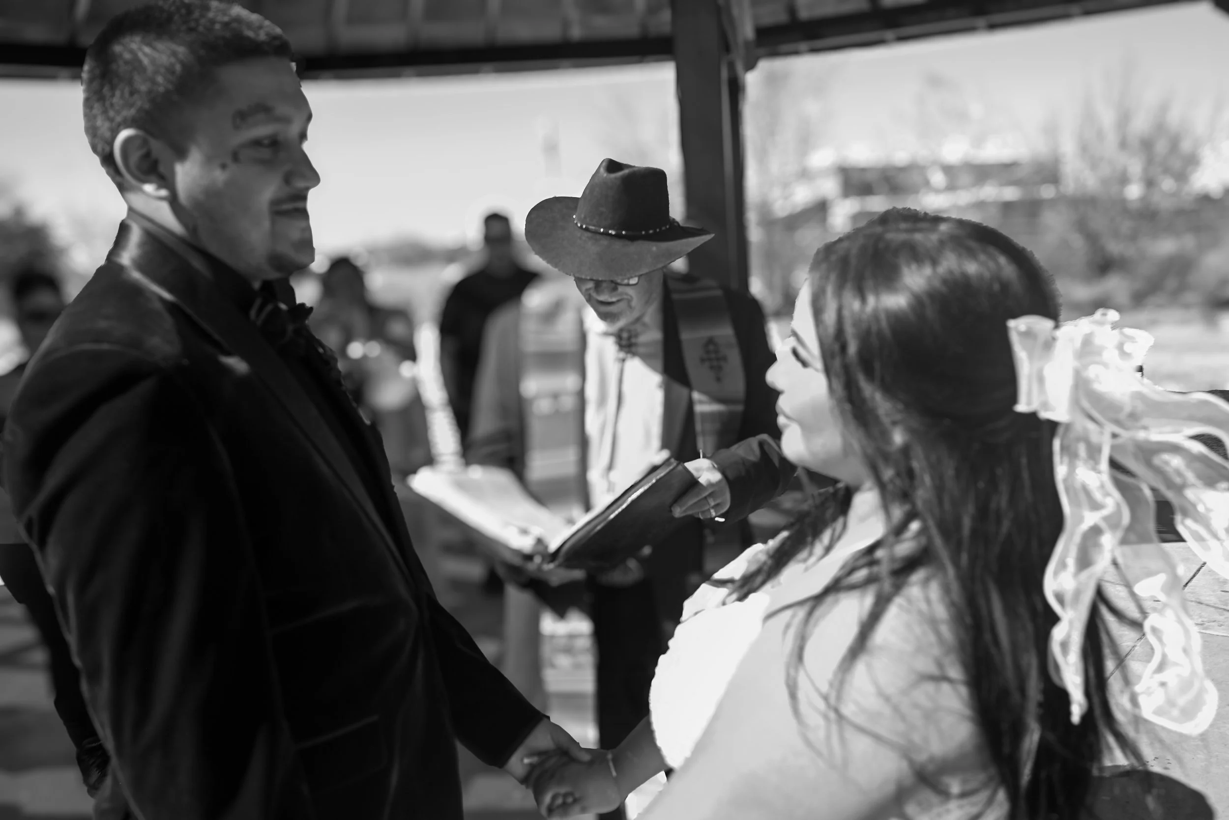 A group of people at an outdoor event, with one person in a tuxedo and others in casual attire, engaging in conversation and looking at a menu or book. A woman in a fancy dress is in the foreground, and a man with a hat and glasses is in the middle.