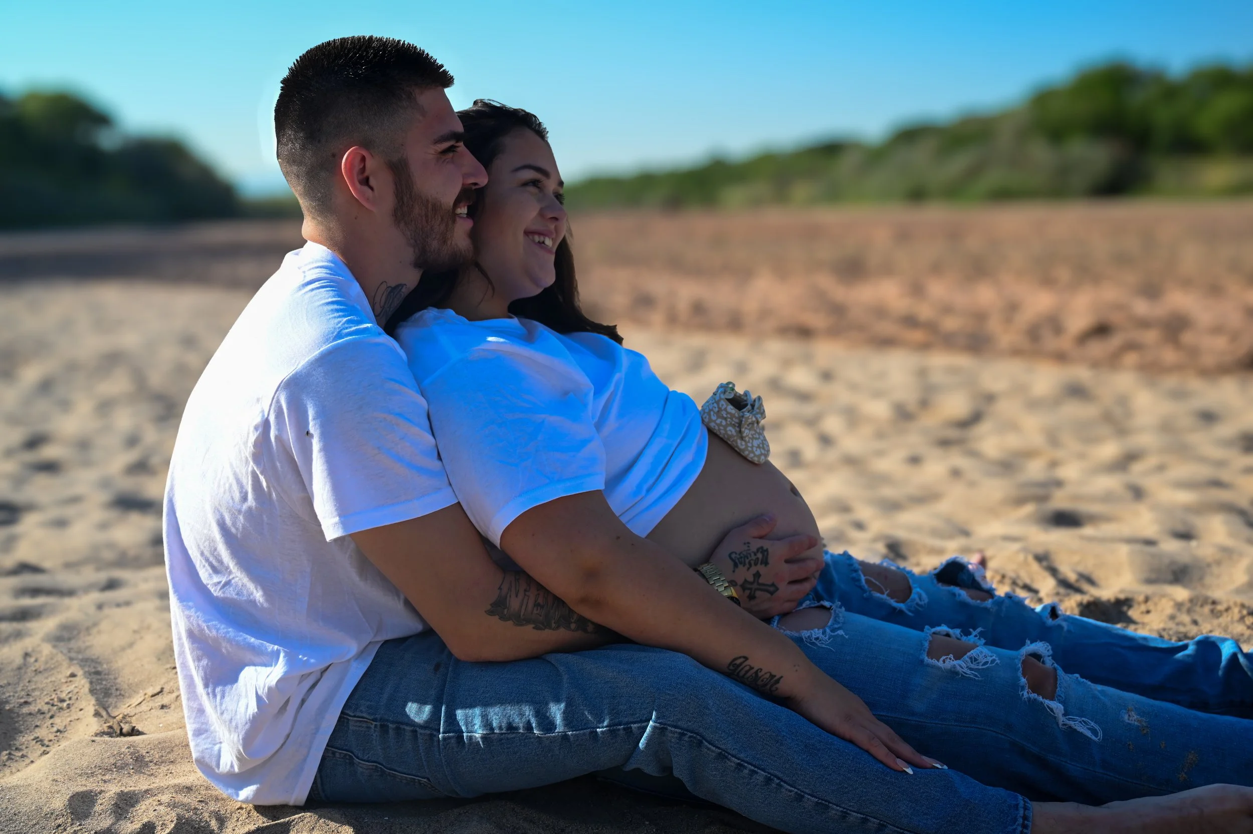 A couple sitting on the beach, hugging and smiling while enjoying the sunny weather and scenic view.