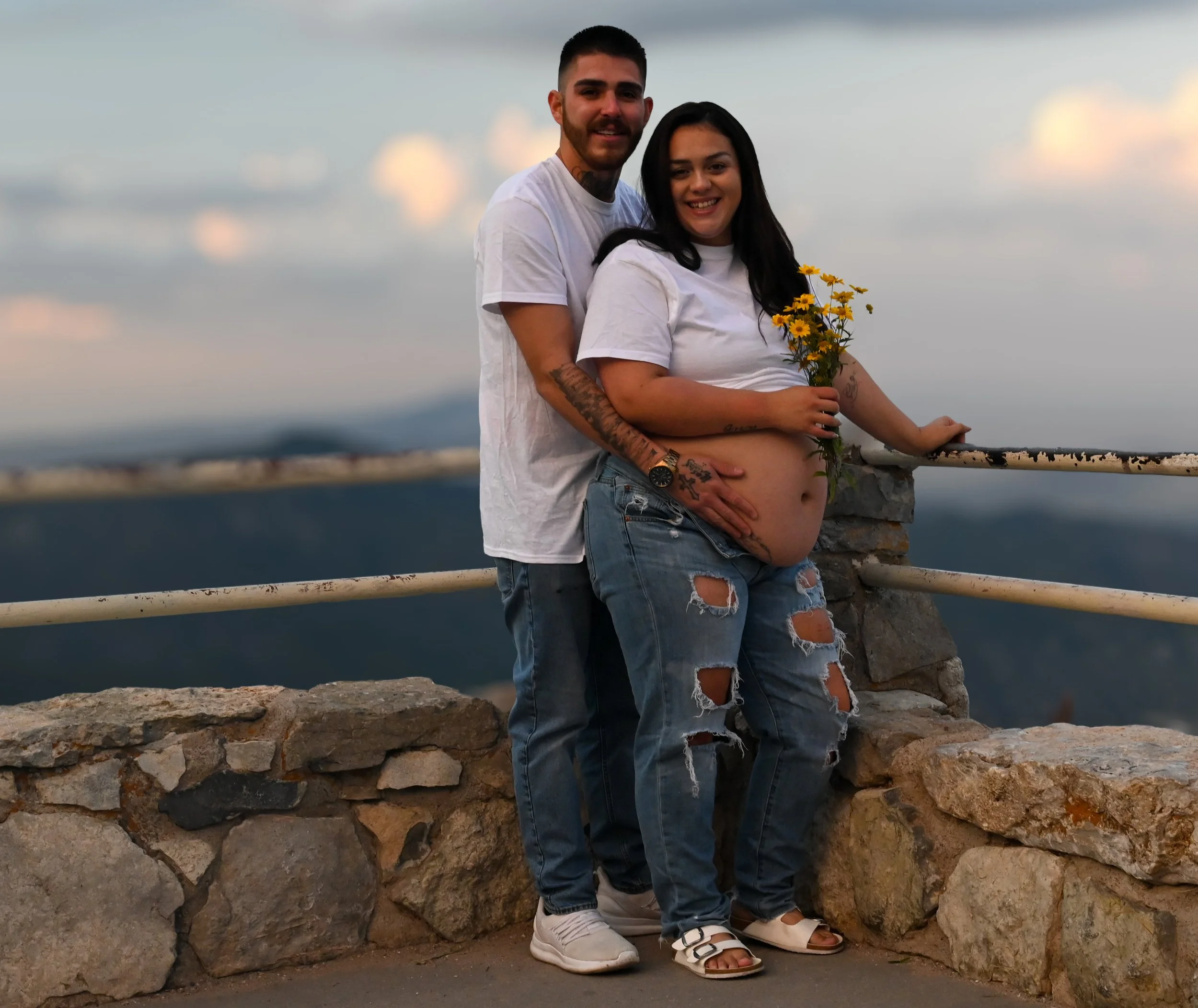 A pregnant woman and a man standing together outdoors on a stone wall, smiling at the camera. The woman is holding a small bouquet of yellow flowers, and she has tattoos on her arms and leg. The man has tattoos on his arm and neck, and they are dress