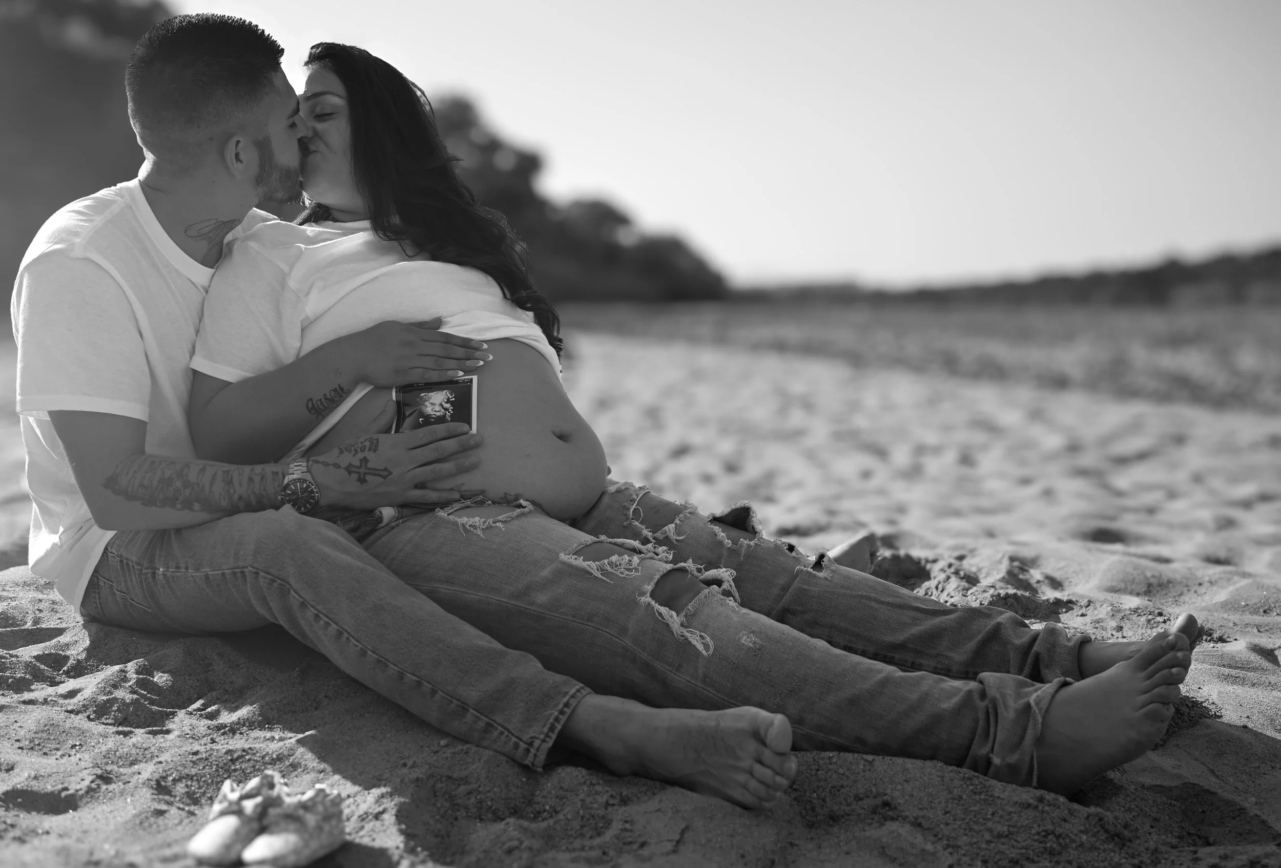 A couple sitting on the beach, embracing and sharing a kiss. The woman is pregnant and holding a photograph of an elderly man. The background shows a sandy beach with a distant shoreline and a clear sky.