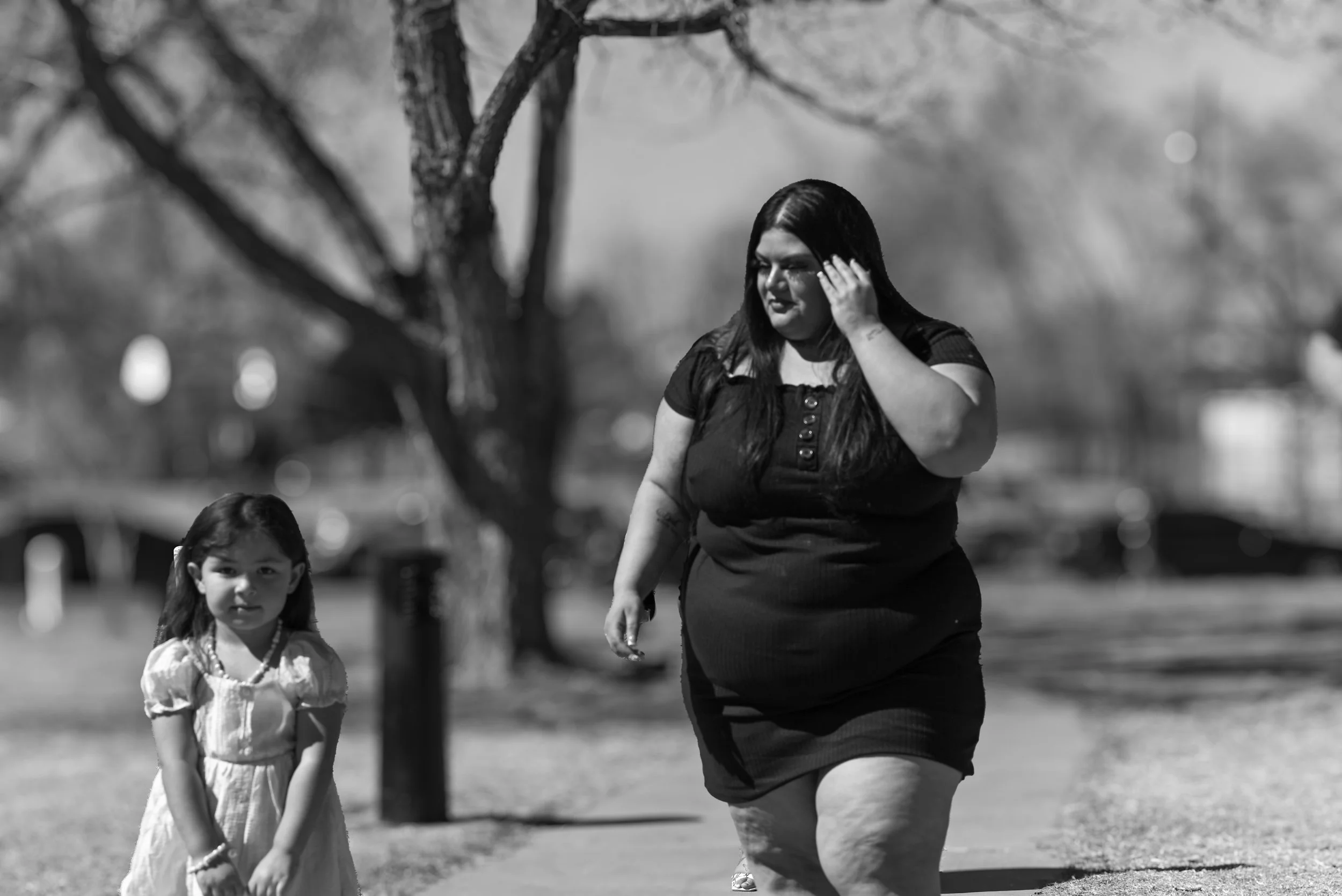 A woman wearing a black dress walks with a young girl in a park with trees and blurred cars in the background, in black and white.