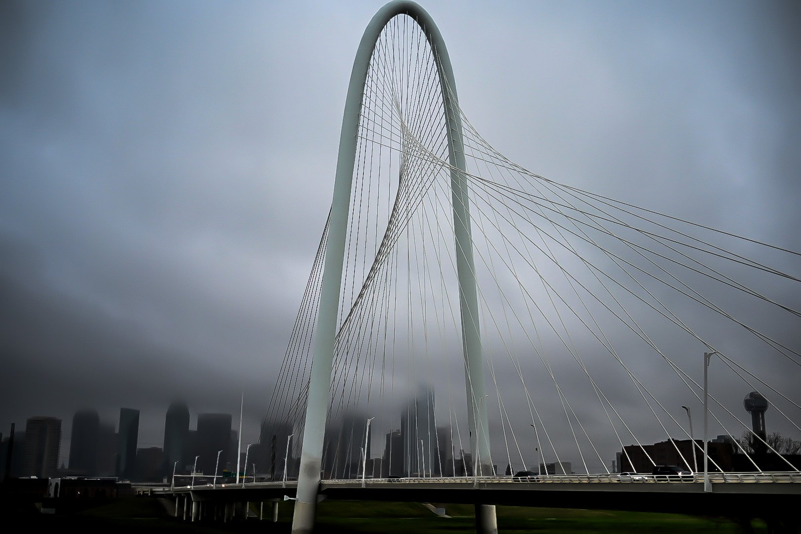 A modern cable-stayed bridge with a large, white, arching support structure under a gray, overcast sky, with a city skyline in the background.