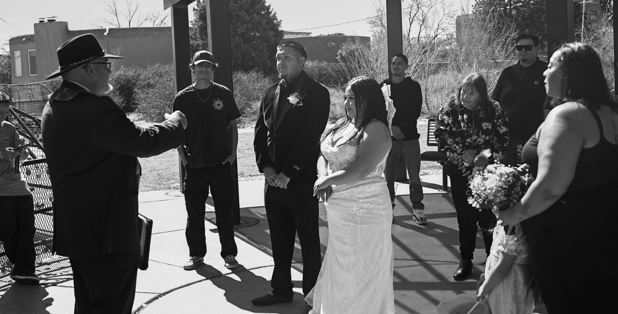 A black and white photo of a wedding ceremony outdoors with guests and the bride and groom standing together, exchanging vows or rings, with an officiant speaking to them.