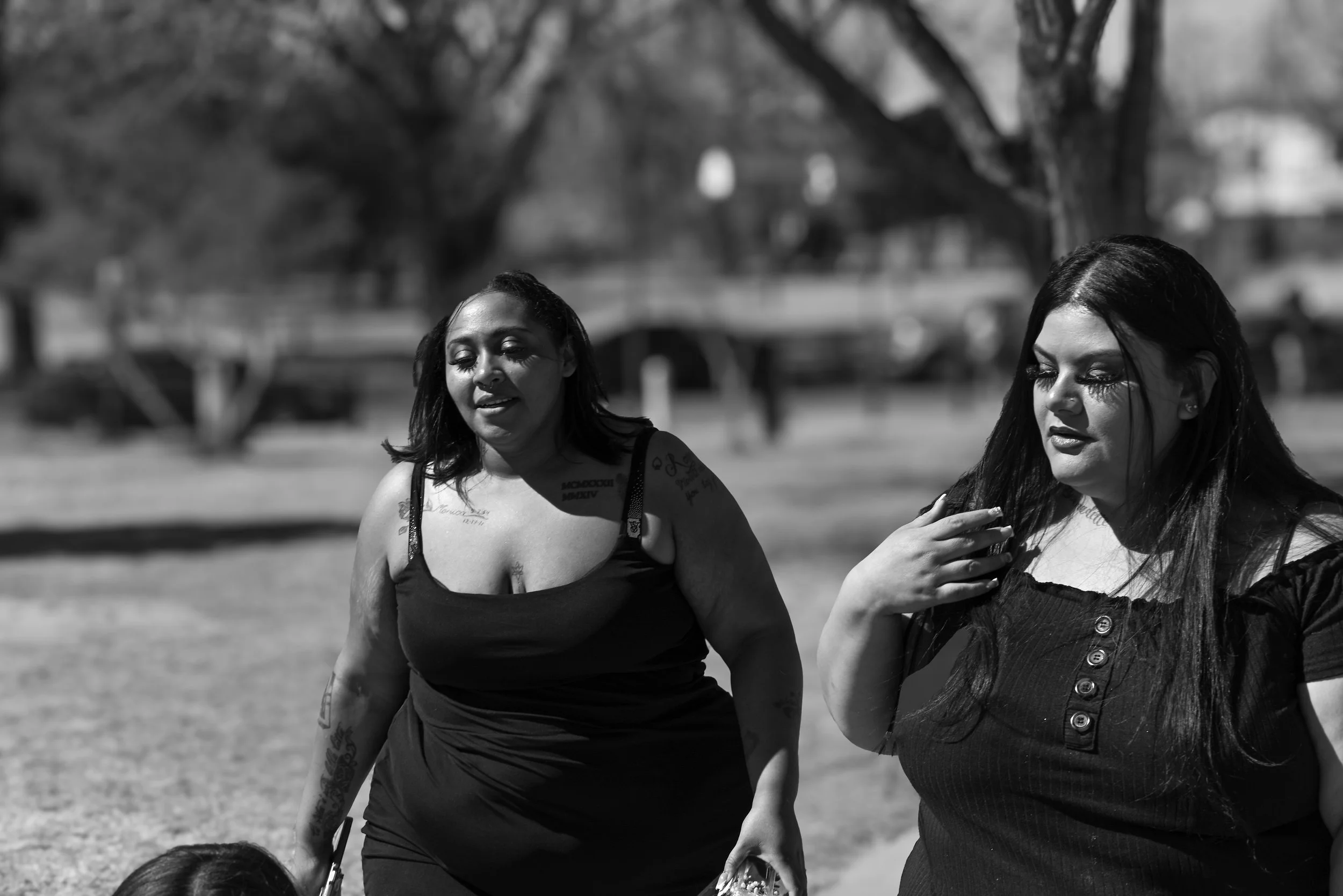 Two women walking outdoors on a sunny day, one with tattoos on her arms and the other with long dark hair and a button-up dress.