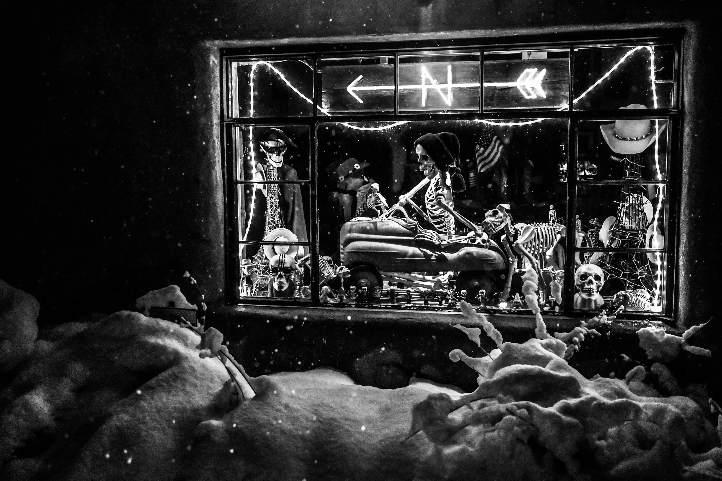 Store window display featuring skeleton figurines, skulls, and Halloween decorations illuminated by neon lights, seen through a snow-covered outside scene.