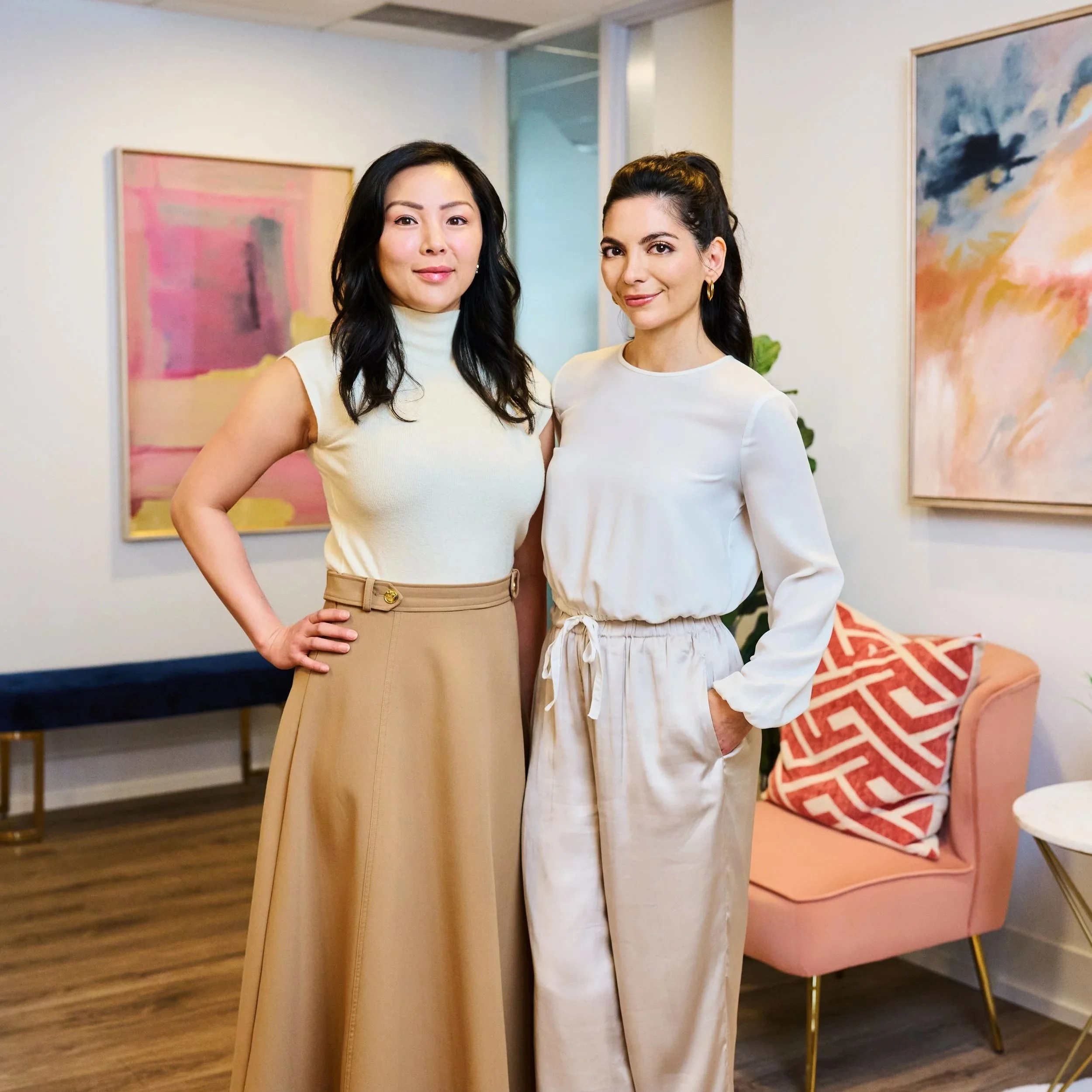 Two women standing in a modern, art-decorated living room. One woman has dark hair, wearing a cream sleeveless turtleneck and tan wide-leg pants with a belt. The other woman has dark hair styled in a half-updo, wearing a white long-sleeve top and matching beige pleated pants. They are smiling and posing for the photo.