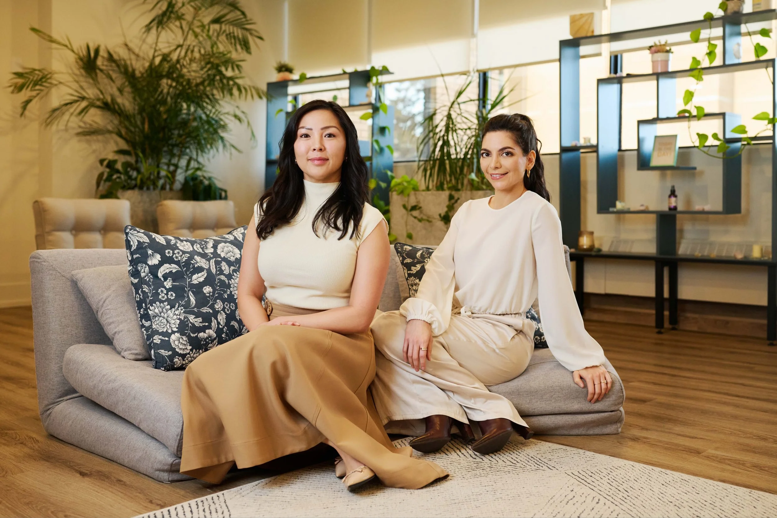 Two women sitting on a light gray sofa with black and white patterned pillows in a modern, well-lit room with wooden flooring and green plants.