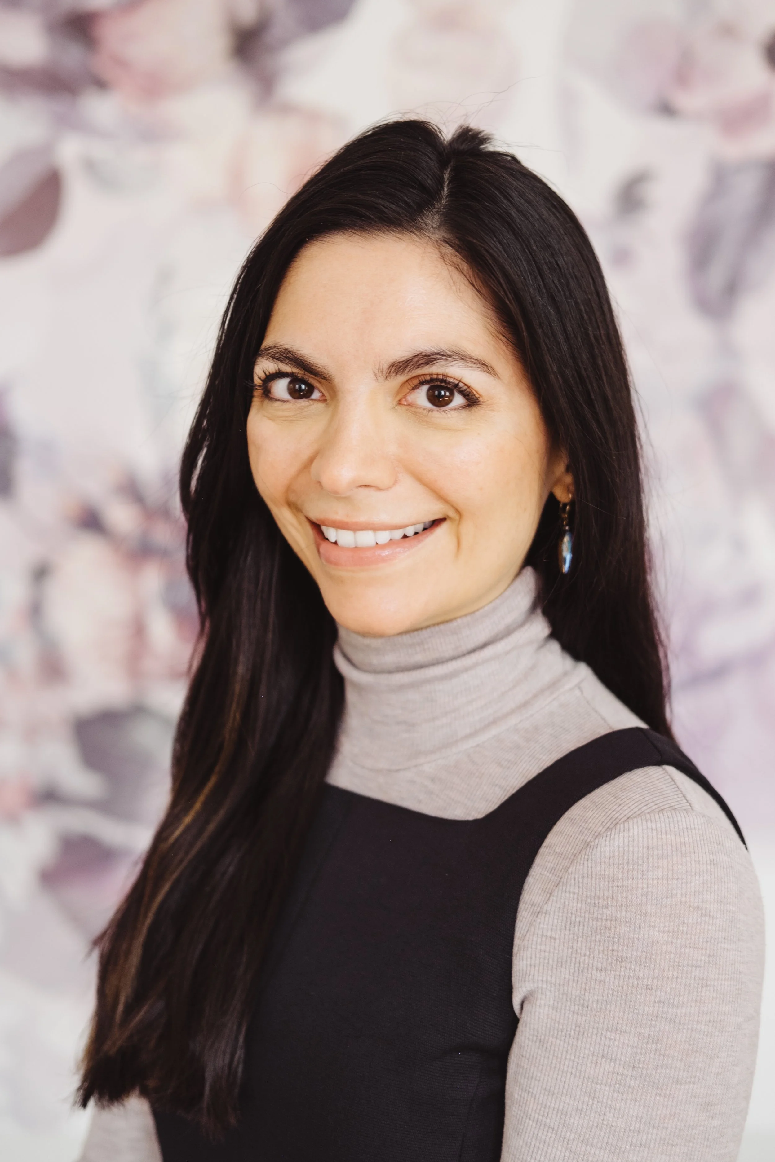 A woman with long dark hair, smiling, wearing a beige turtleneck and a black sleeveless dress, with a light-colored floral background.