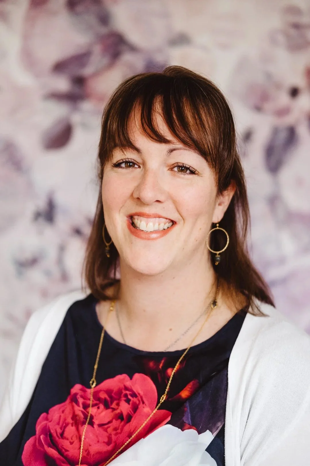 A woman with shoulder-length brown hair and bangs, smiling and showing her teeth, wearing hoop earrings and a necklace, against a blurry floral background.