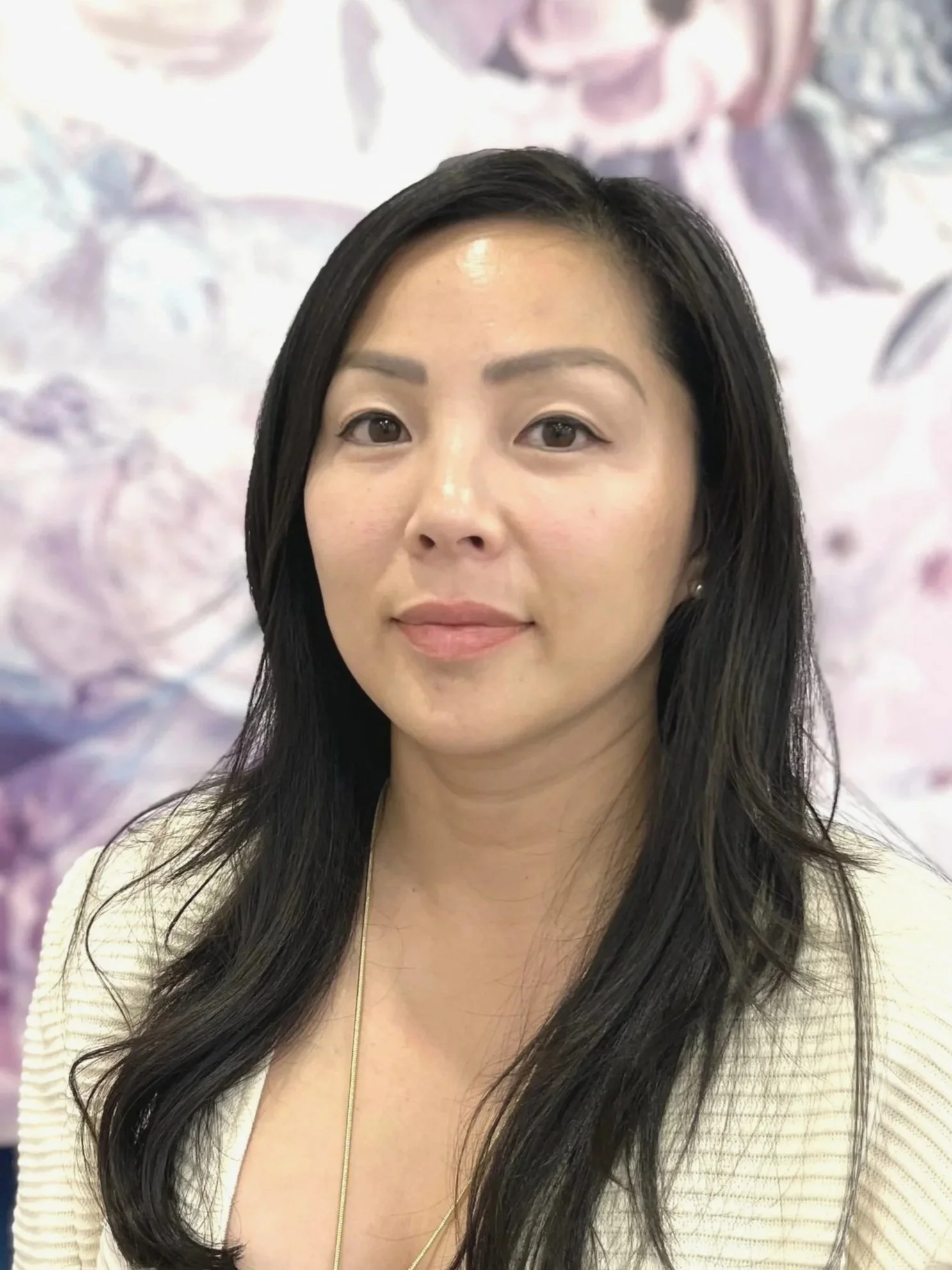 A woman with long black hair and a light-colored top looking at the camera with a subtle smile, standing in front of a blurred pink and purple floral background.