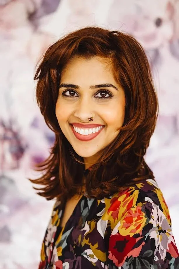 Smiling woman with shoulder-length brown hair, wearing a colorful floral top, standing in front of a soft pink and purple blurred background.