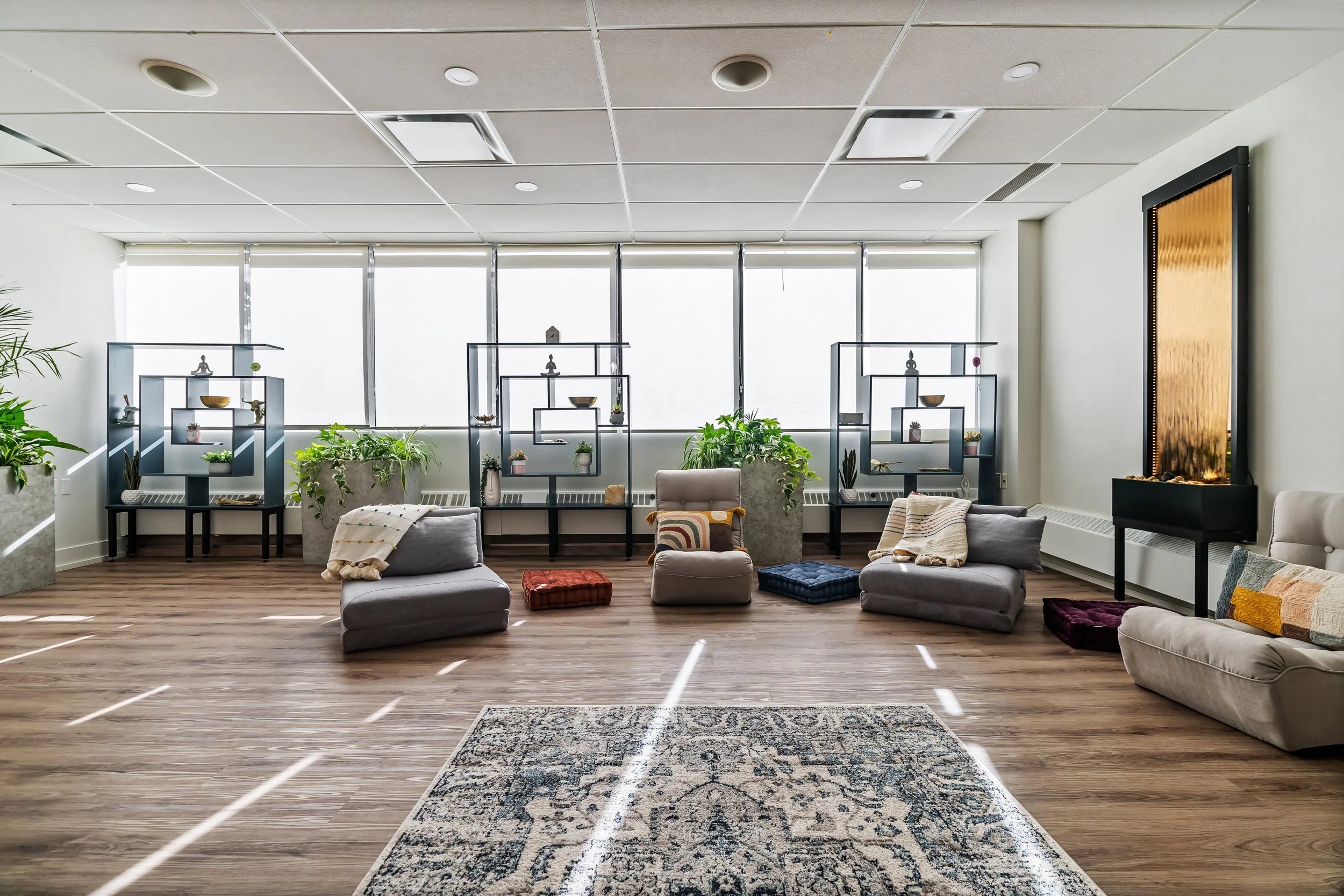 Modern lounge with gray couches, colorful cushions, indoor plants, decorative shelves, and a water feature against large windows with blinds, wooden flooring, and a patterned area rug.