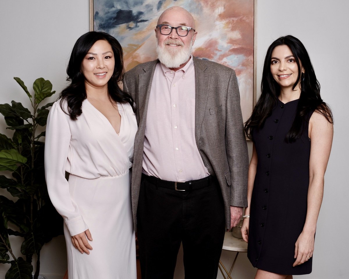 Three people standing together indoors, posing for a photo, with a colorful abstract painting behind them and green plant on the side.